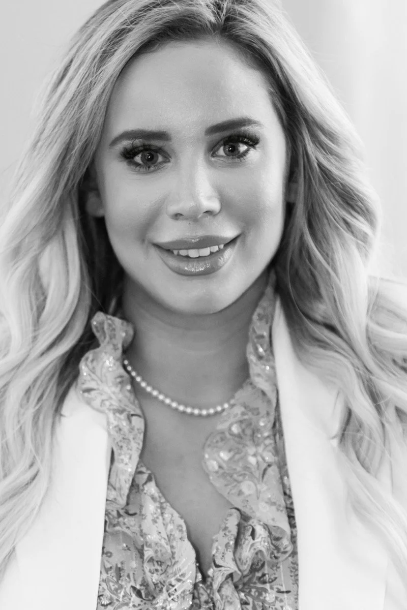 Black and white portrait of a smiling woman with long, wavy hair, wearing a pearl necklace and a patterned blouse, with a blazer over it.