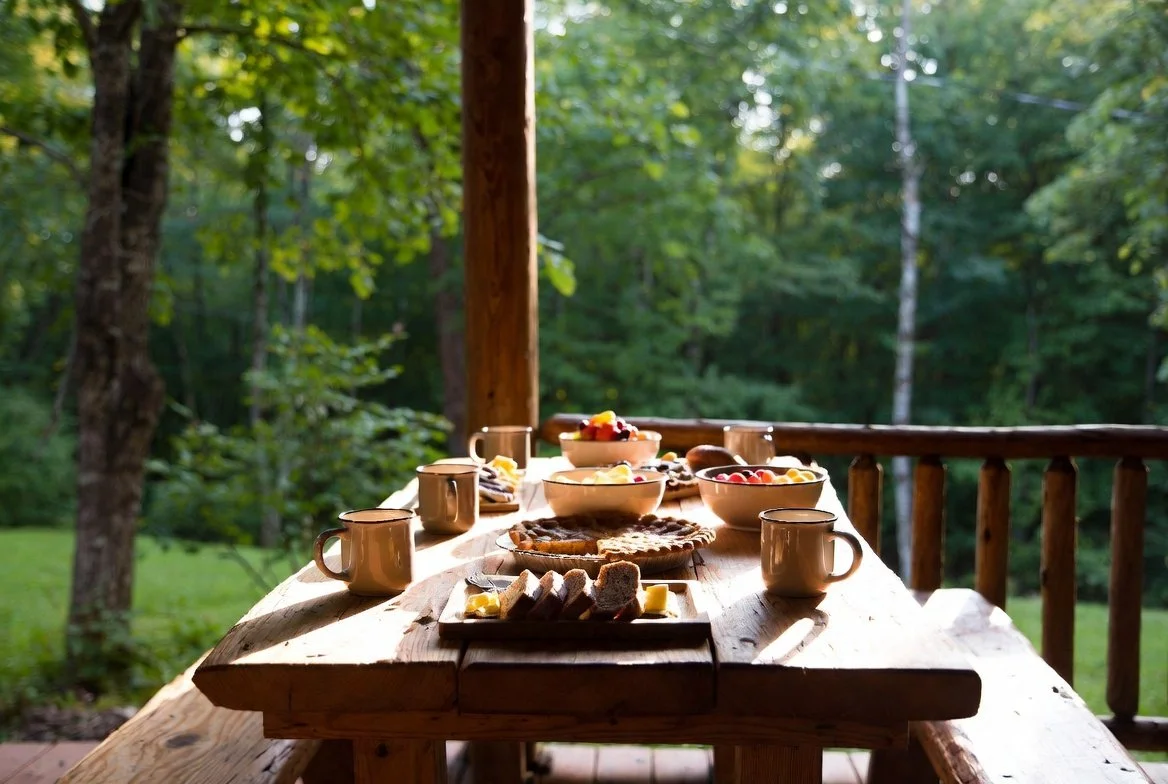 Outdoor breakfast table with various food items on a wooden deck surrounded by trees