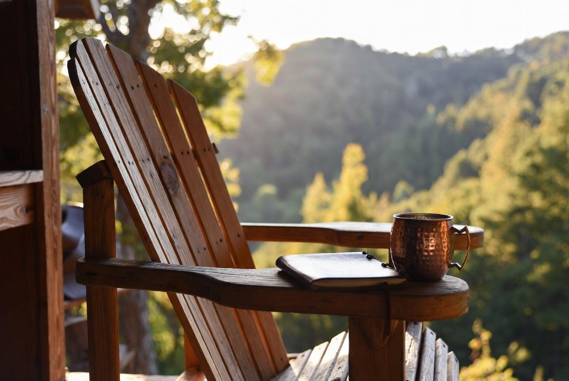 Wooden adirondack chair on a porch facing a forested landscape with mountains in the background, with a copper mug and a notebook on the armrest.