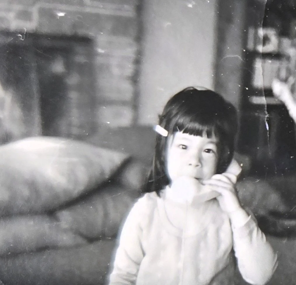 A young girl--child that looks around 4 years old with almost shoulder-length black hair, wearing a light-colored top. She's looking at the camera talking on what looks like an old-school toy phone in a black & white photo. Presumably Frannie James.