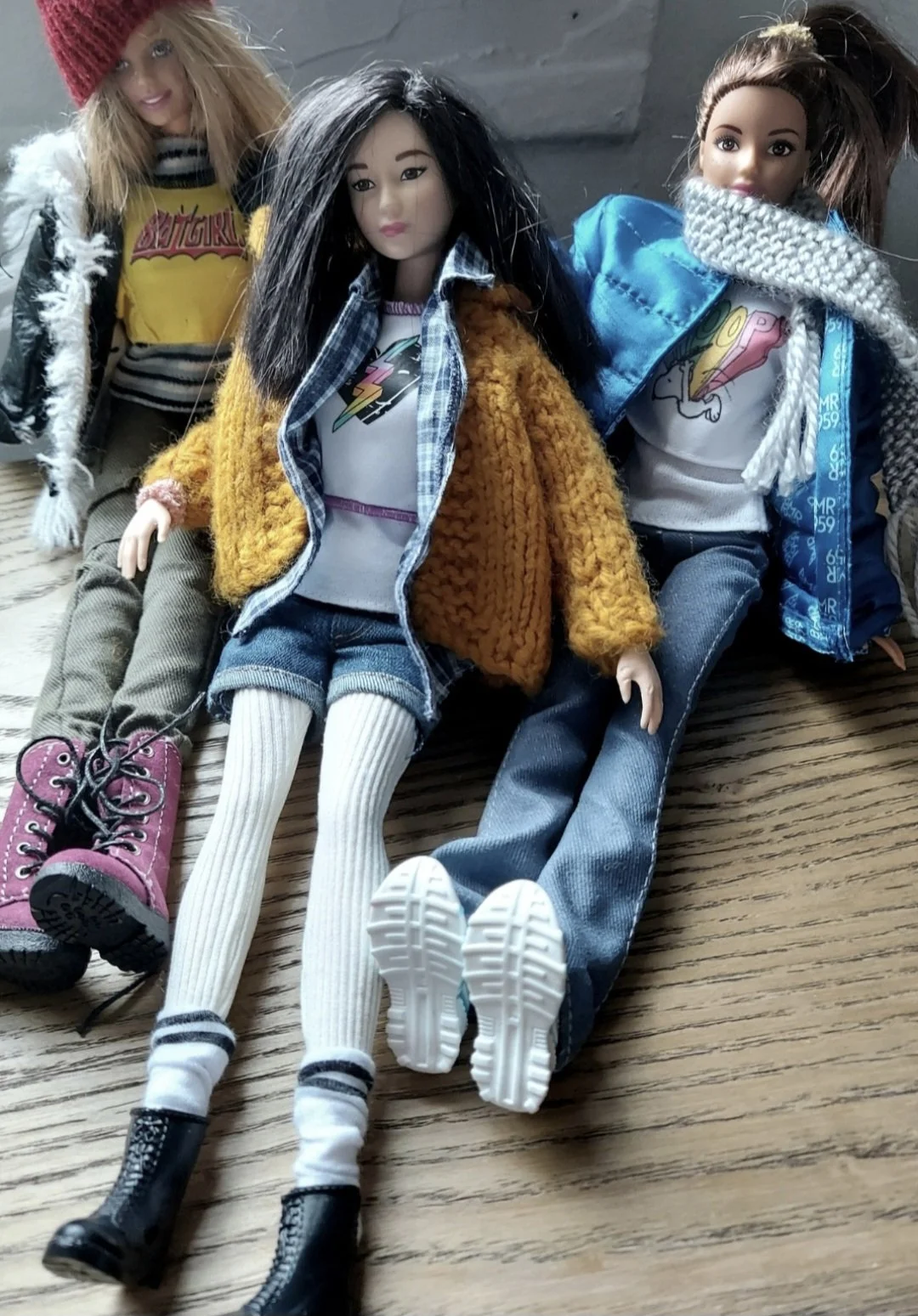 Three Barbie dolls, each dressed in 90s clothing, propped on a wooden surface, sitting together. The dolls are wearing various jackets, shirts, pants, and boots with different styles and colors.