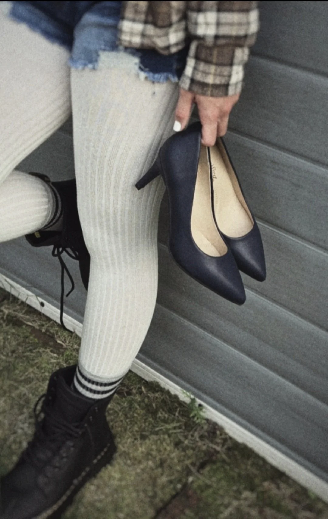 Photo of a girl from the waist down, standing against a wall and dressed in 90s-Seattle-style clothing including a flannel shirt, white ribbed tights under short denim cutoff shorts, black combat boots. She's also holding a pair of navy high heels.
