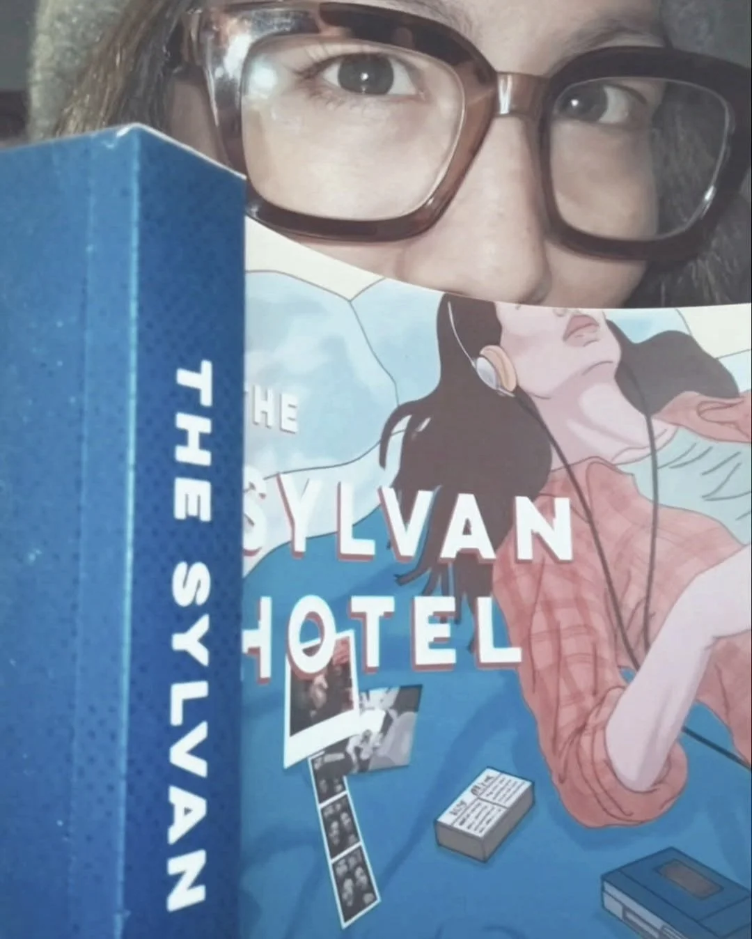 A woman wearing glasses partially hiding her face behind a blue box labeled 'THE SYLVAN HOTEL' and a comic-style illustrated book cover showing a woman with headphones.