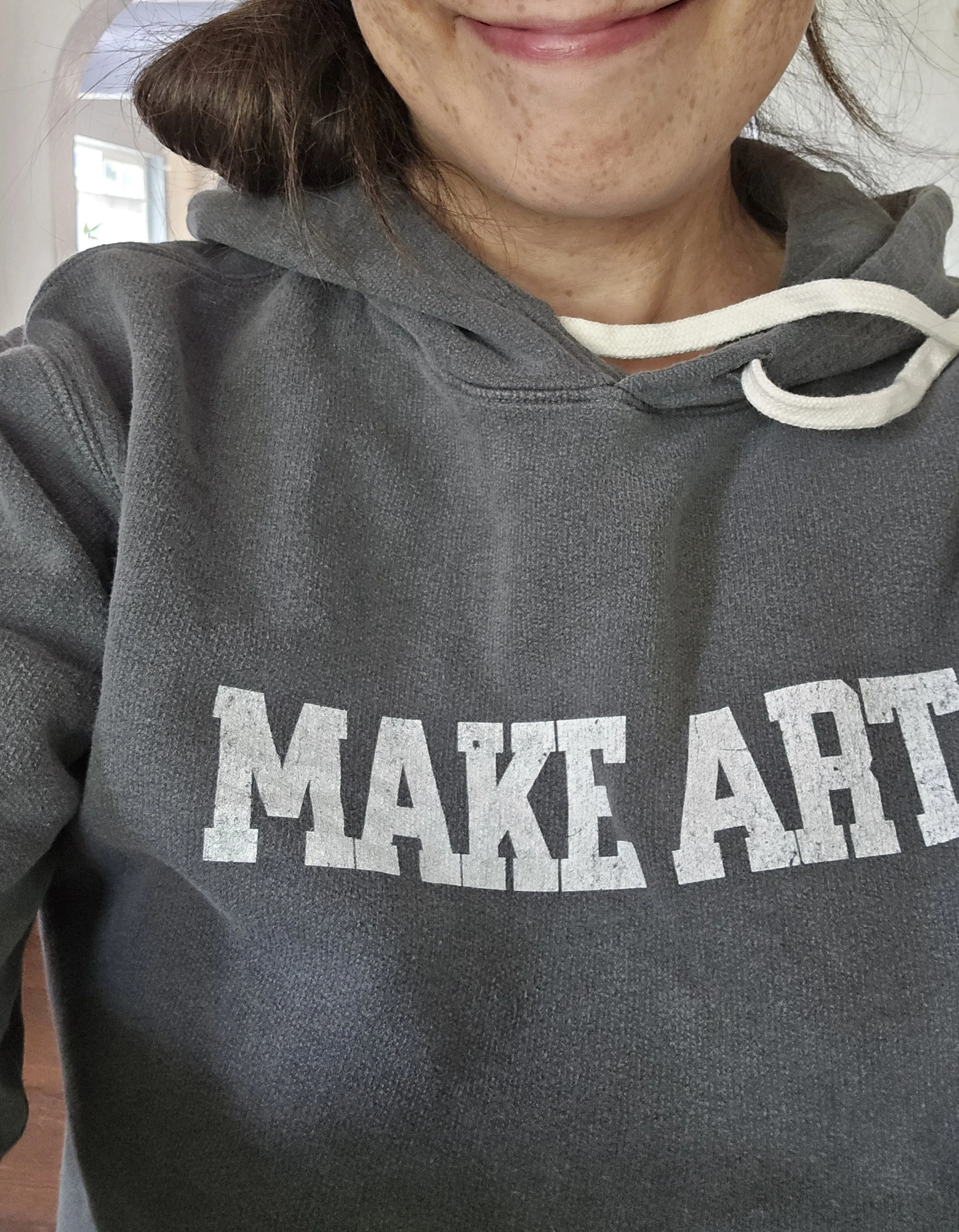 Close-up of a person in a gray hoodie with white drawstrings, with part of their face and smile visible. The hoodie has white text on it that reads "MAKE ART." Image is next to the the "About Frannie James" section so presumbably it's Frannie James.