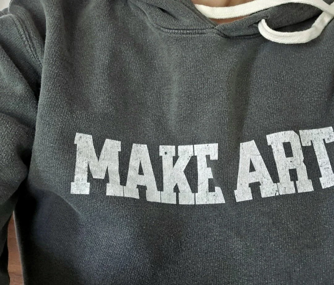 Close-up of a person wearing a gray hoodie with the words 'MAKE ART' printed in large white letters on the front.