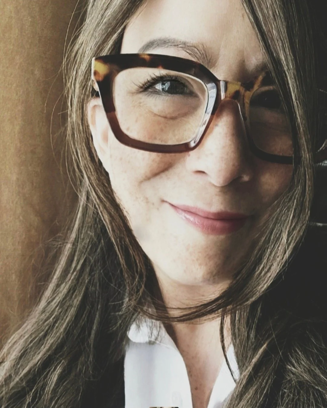Close-up of a woman with long brown hair and wearing large tortoiseshell glasses, smiling slightly.