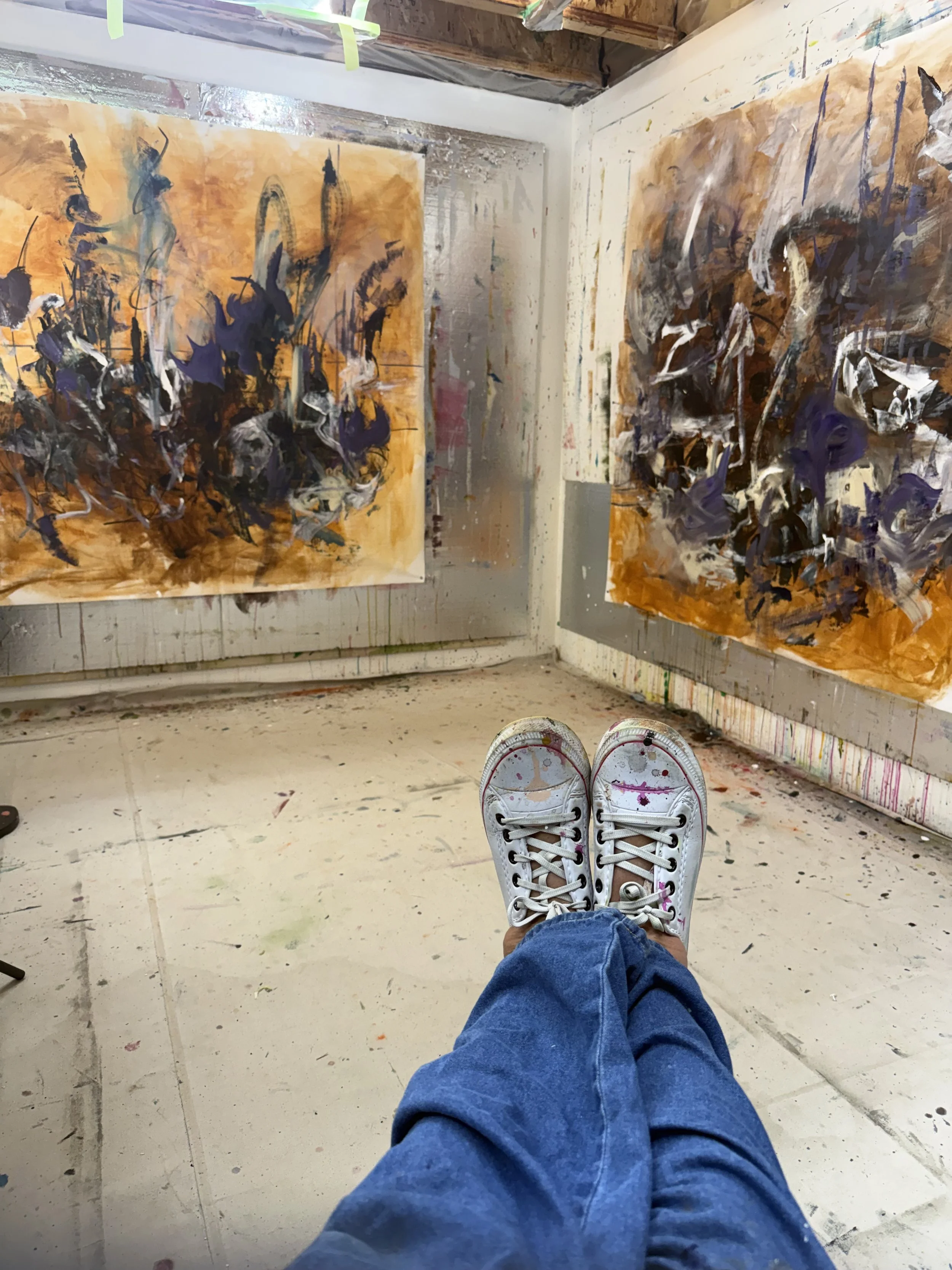 A person sitting with legs outstretched, wearing white sneakers splattered with paint, in an artist's studio with abstract paintings on the walls.