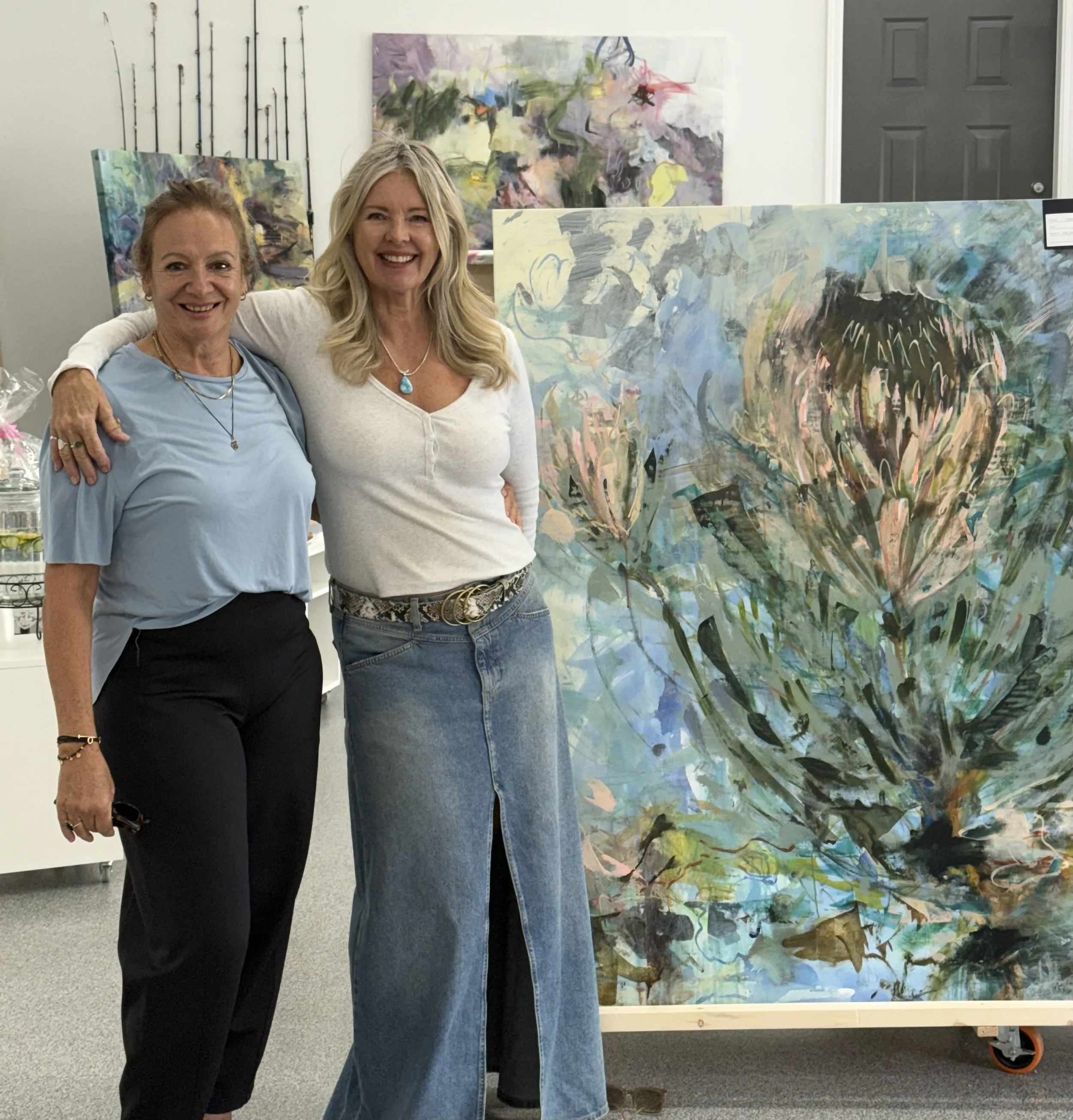 Two women standing next to each other in front of colorful abstract paintings, smiling, with one woman having her arm around the other's shoulder.