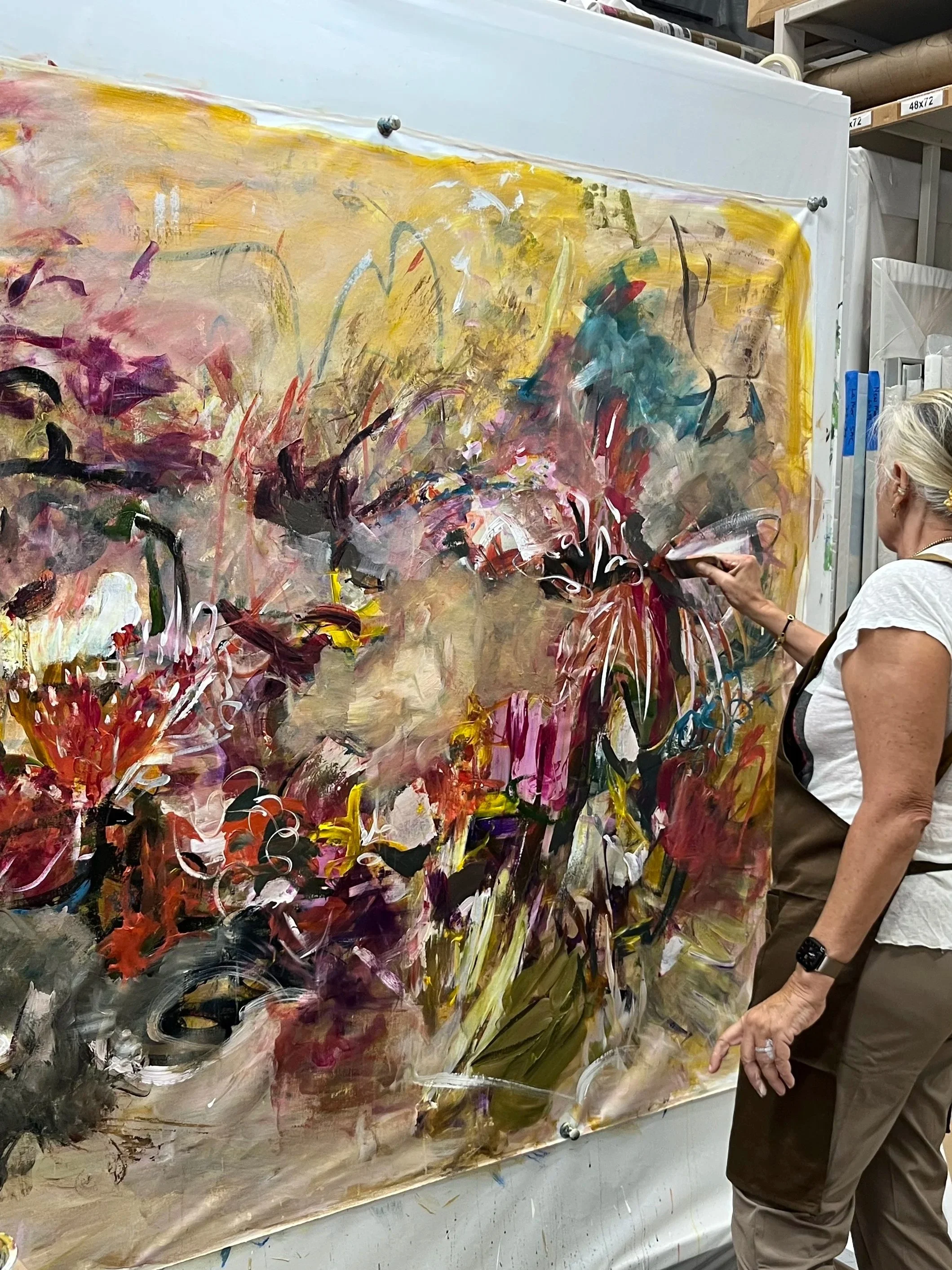 A woman with gray hair and wearing a white shirt and khaki pants is painting a large abstract expressionist artwork on canvas with various colors, including yellow, red, purple, black, and white, in an art studio or gallery setting.
