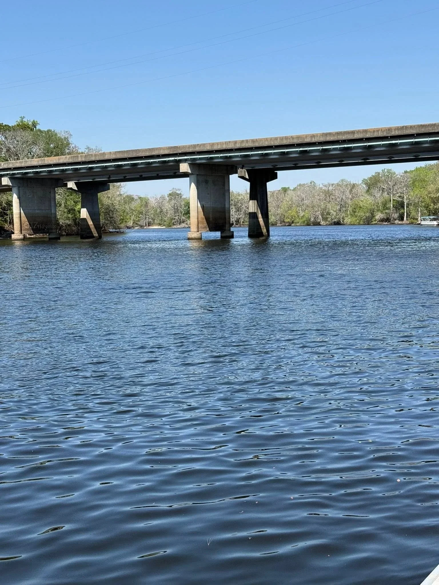 Water body with gentle ripples and a bridge crossing over it, with trees in the background under a clear blue sky.