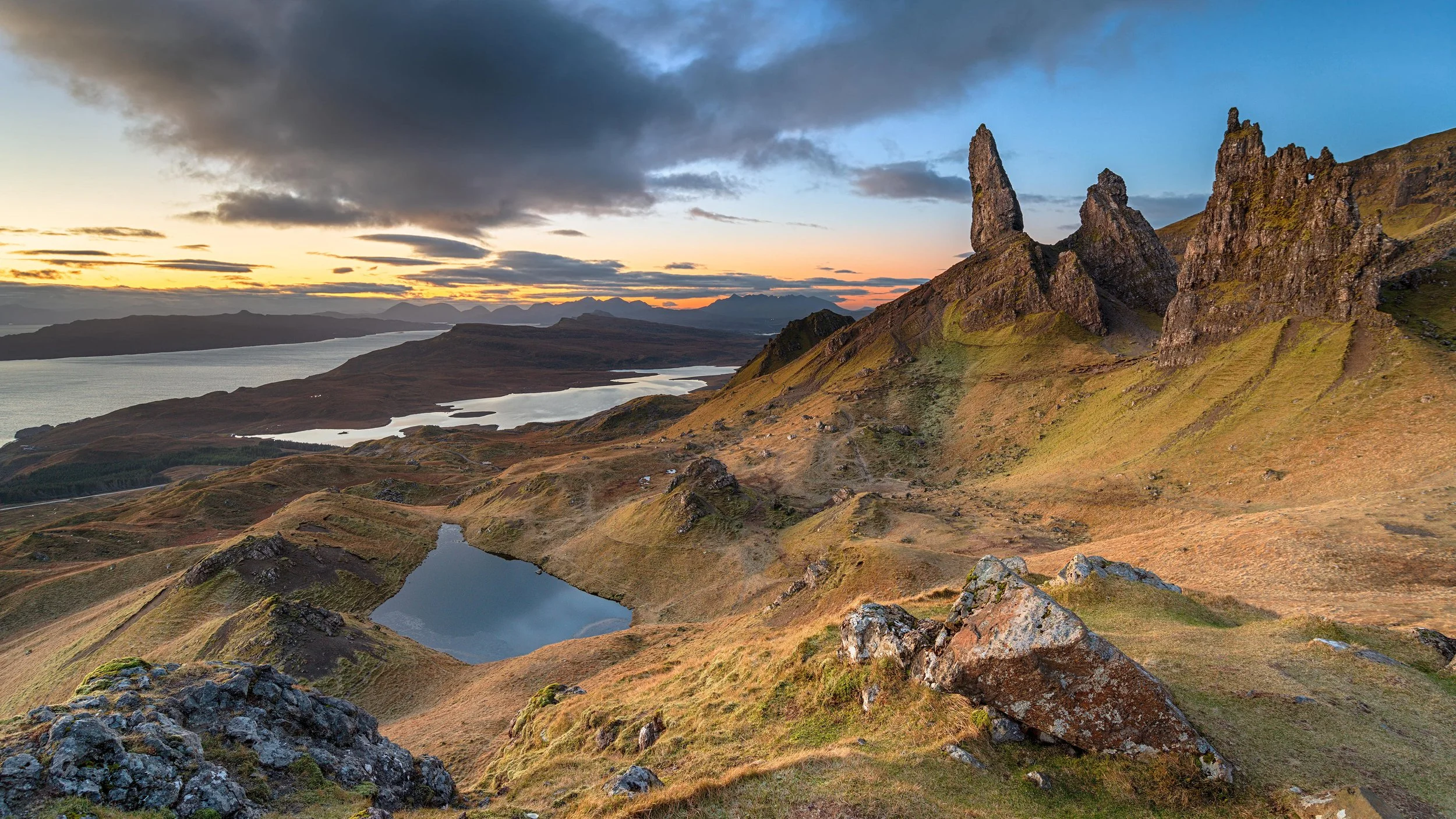 Scenic landscape of rolling hills, several lakes, and rock formations with a sunset sky and dark clouds in the background.