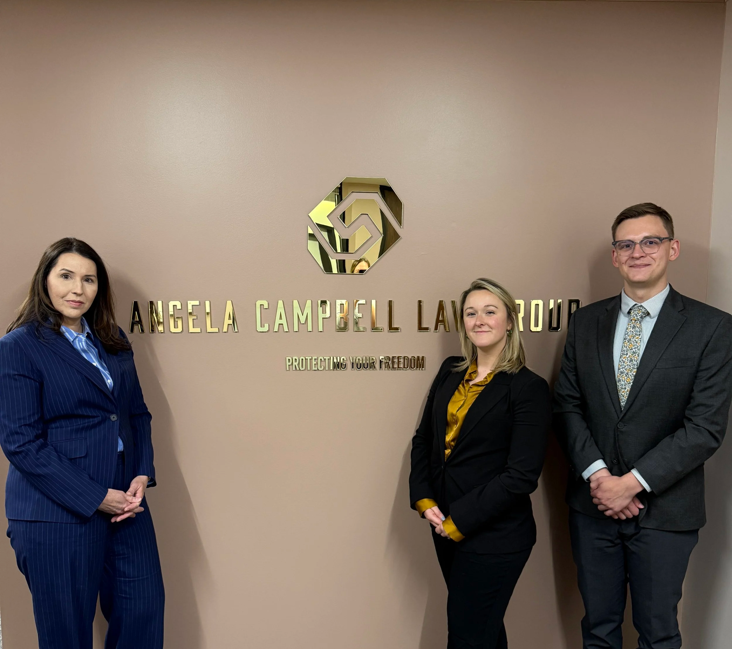 Four individuals standing in front of a beige wall with a metallic logo and gold text that reads "Angela Campbell Law Group." The group includes two women and two men, all dressed professionally, smiling at the camera.