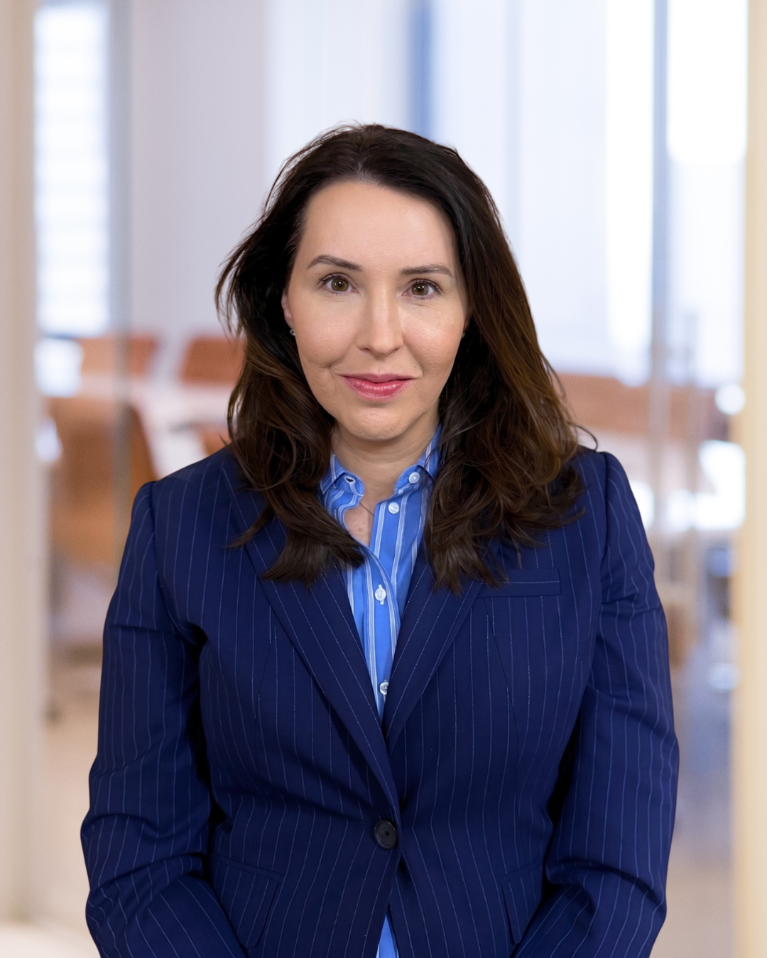 A woman with dark brown hair wearing a blue pinstripe blazer and a blue collared shirt, standing in a professional office setting with a blurred background of a meeting room.