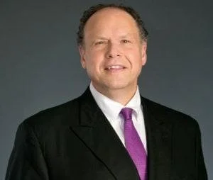A man wearing a black suit, white shirt, and purple tie against a gray background.
