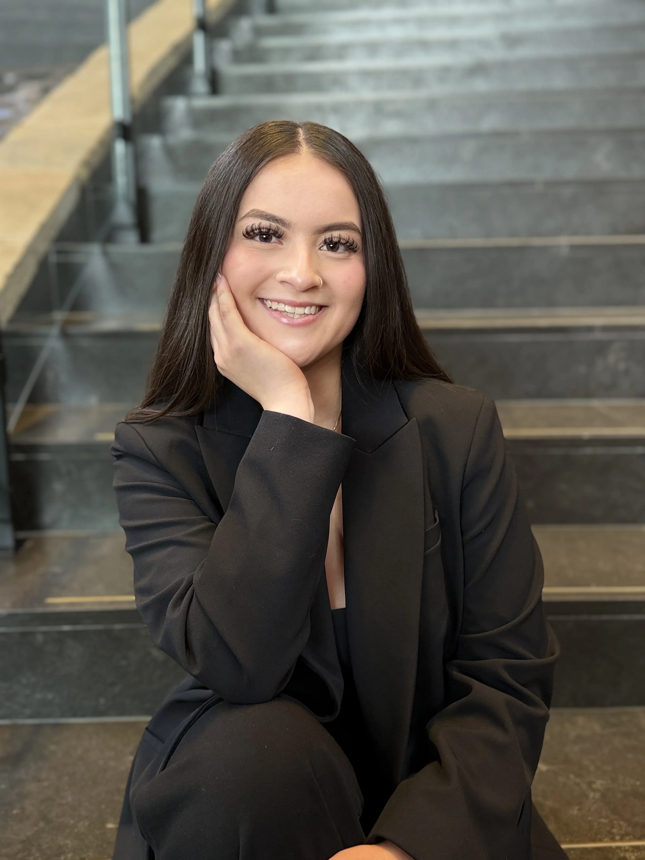 A young woman with long dark hair, wearing a black blazer, smiling, sitting on stairs.