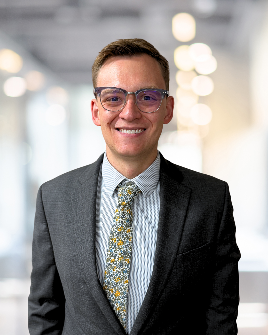 A smiling young man wearing glasses, a dark suit, a striped shirt, and a floral tie, standing in a well-lit, modern office corridor.
