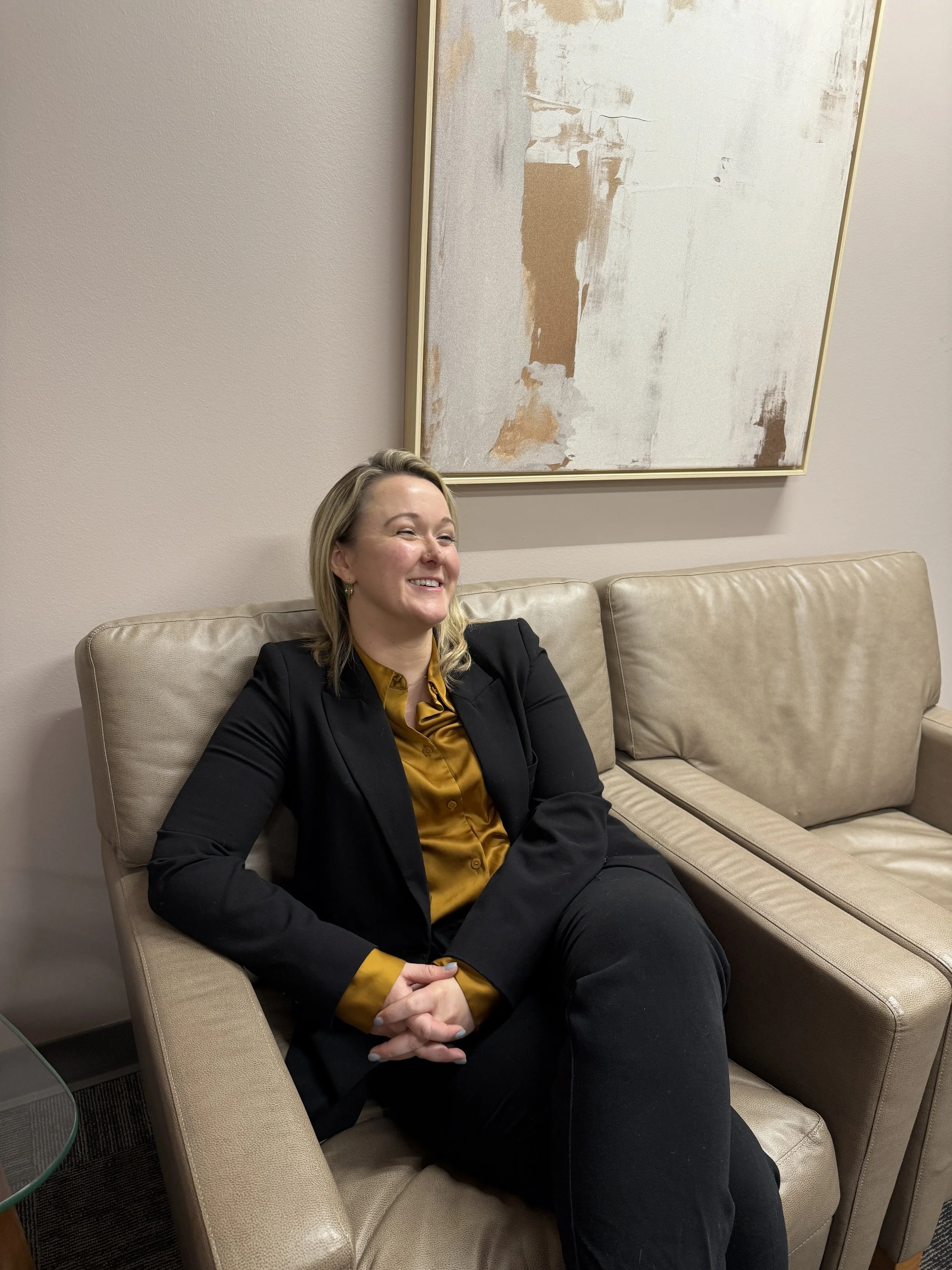 A woman with blonde hair sitting on a beige sofa, smiling, wearing a black blazer, mustard yellow shirt, and black pants, with a large abstract painting hanging on the wall behind her.