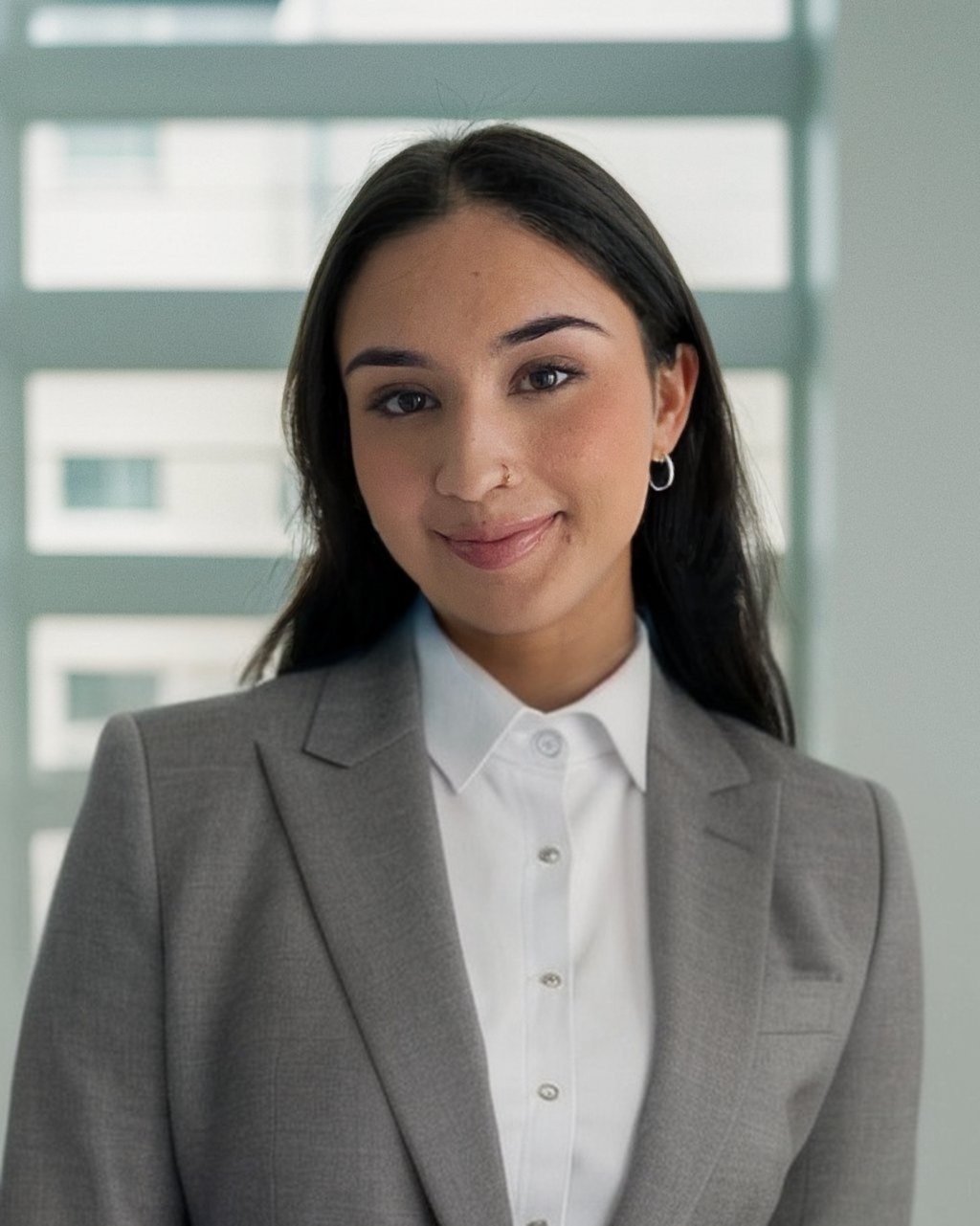 A young woman with dark hair, wearing a gray blazer and white shirt, smiling in front of a modern window background.