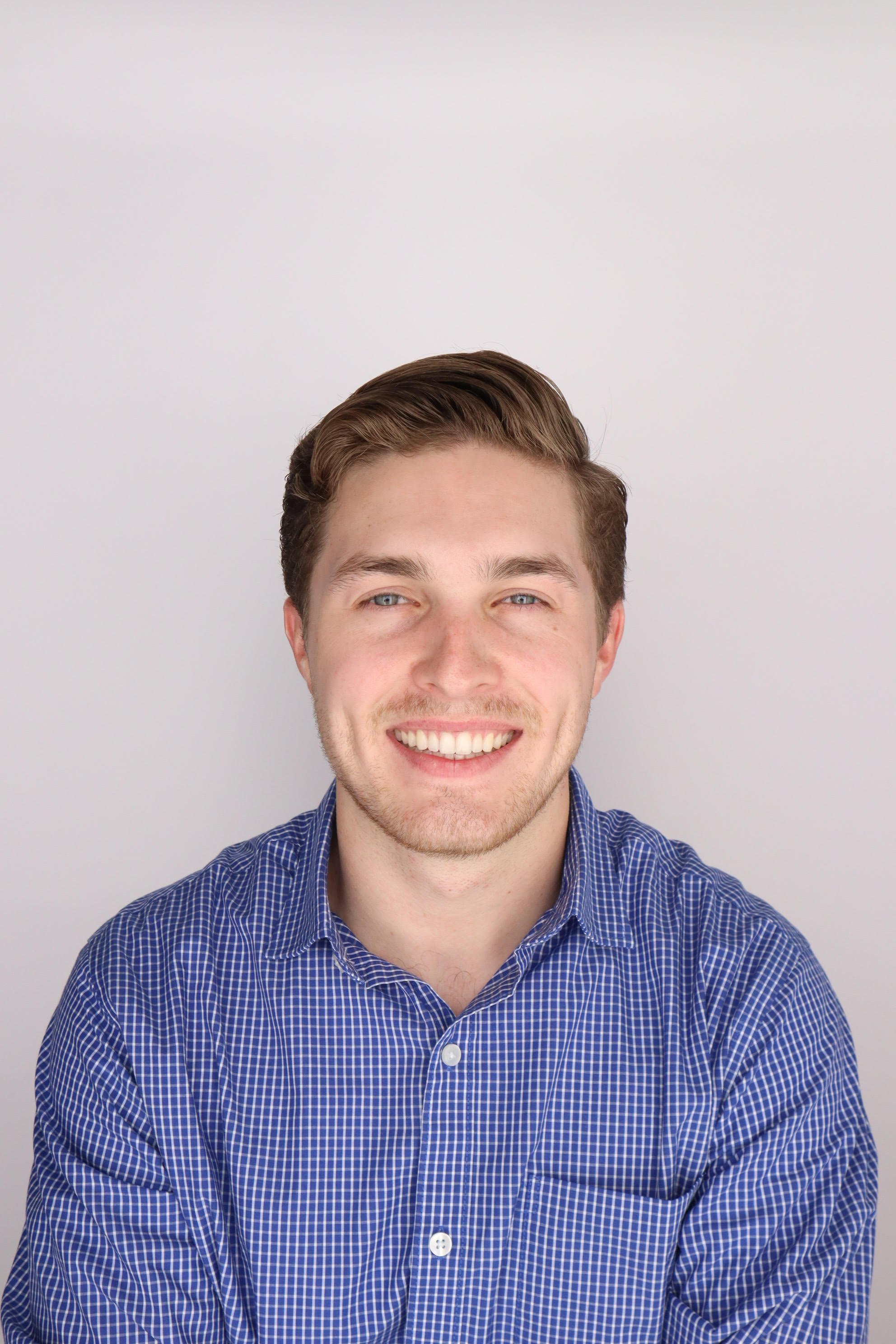 Smiling young man with brown hair and blue eyes, wearing a blue checkered shirt, against a plain light background.