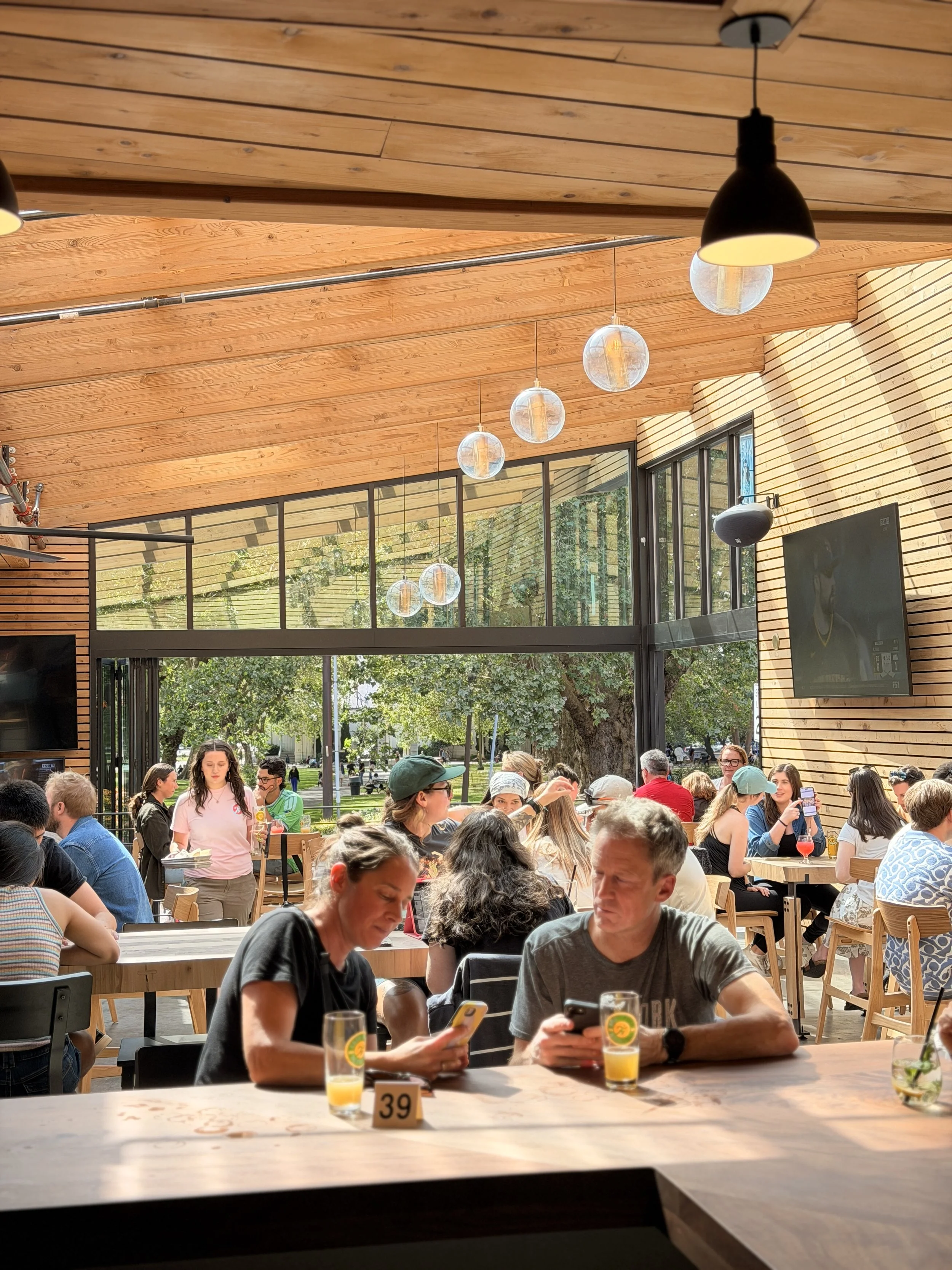 People dining at a modern restaurant with wooden ceilings, large windows, and hanging light fixtures, with outdoor greenery visible outside.