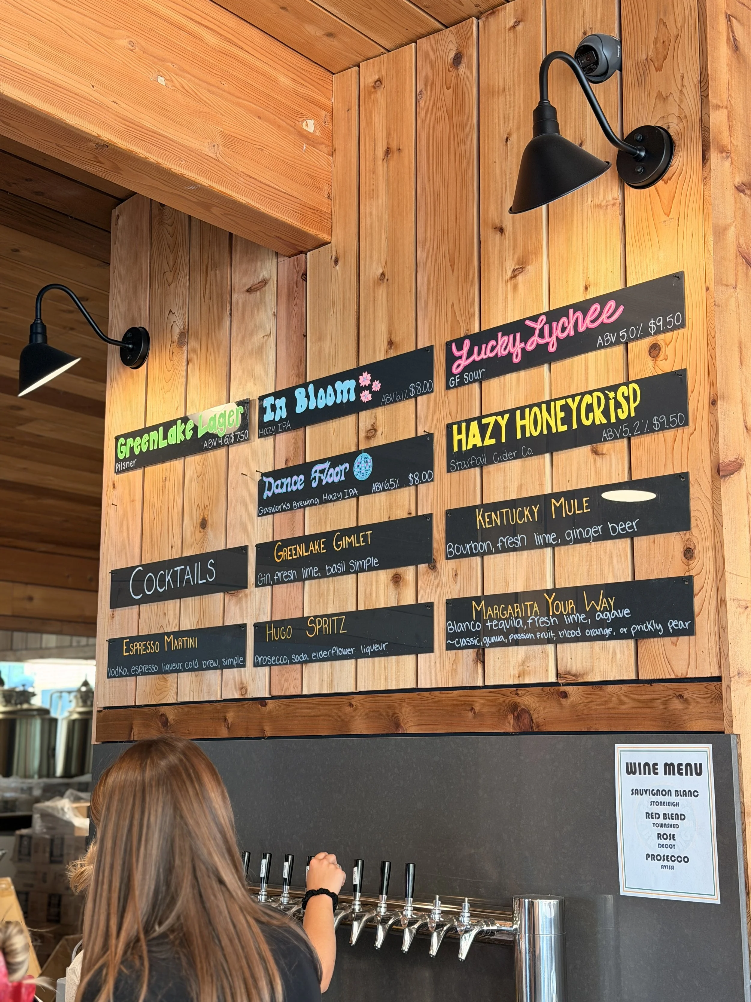 Person with long brown hair pouring a drink at a bar with black wall-mounted beer taps. Behind the person, there are chalkboard-style menus listing various cocktails and their ingredients, as well as a wine menu on a white sheet of paper on the right side of the wall.