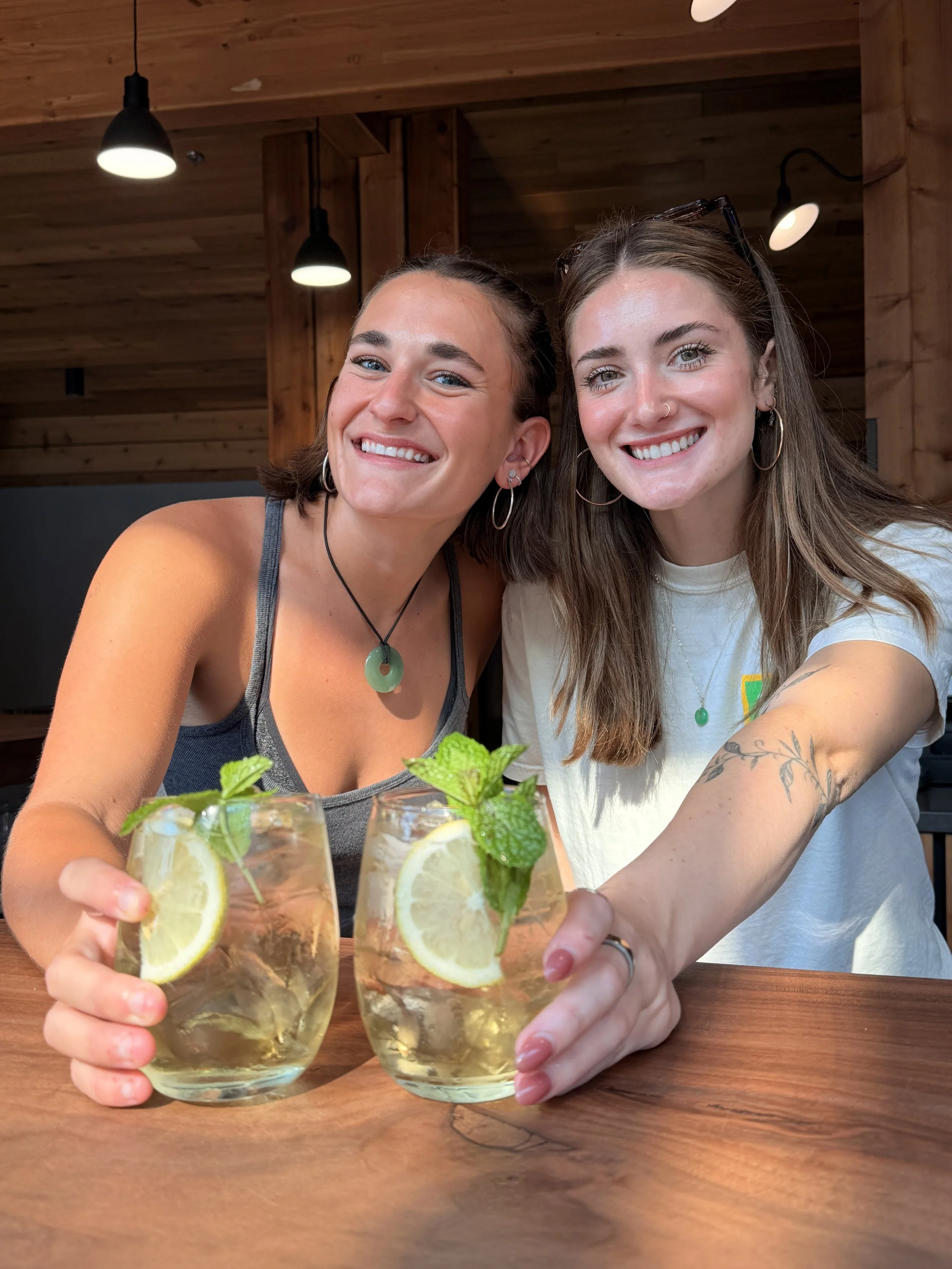 Two women smiling and clinking glasses of lemonade with lemon slices and mint leaves, seated at a wooden table in a cozy, wood-paneled room.