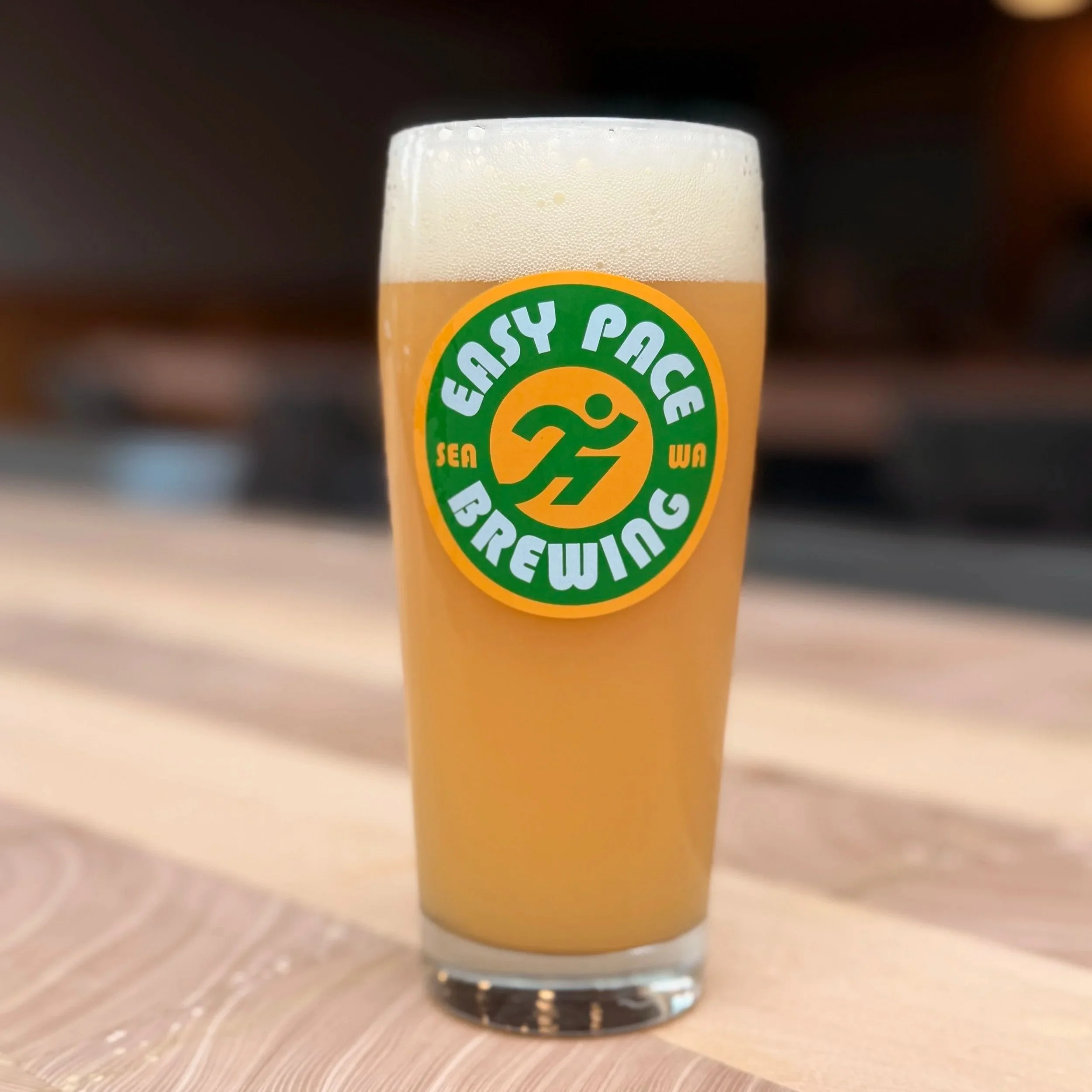 Close-up of a glass filled with a hazy, pale yellow beer, topped with white foam, placed on a light-colored wooden surface.