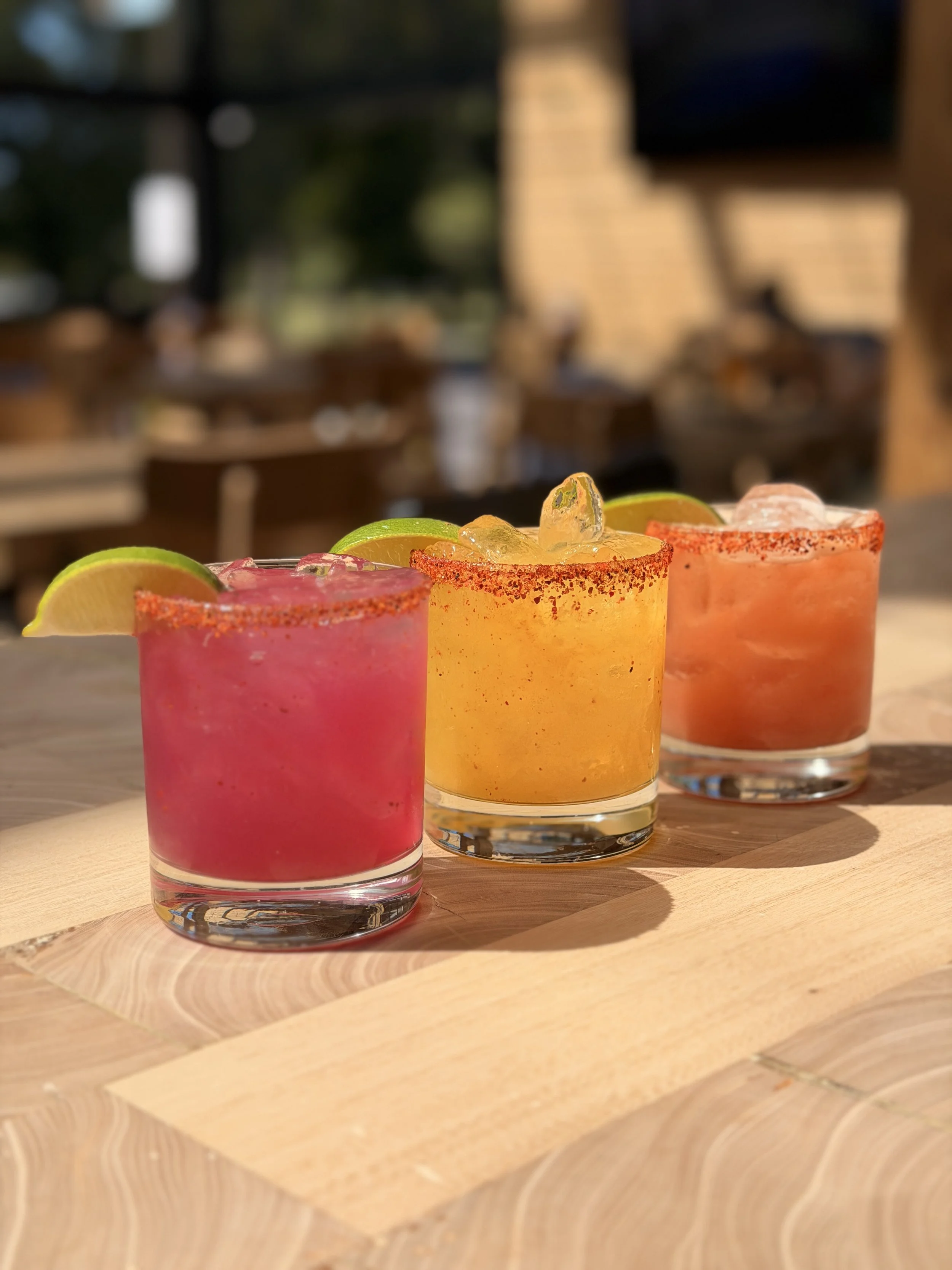 Three colorful cocktails with lime wedges and salt rims on a wooden table, blurred background.