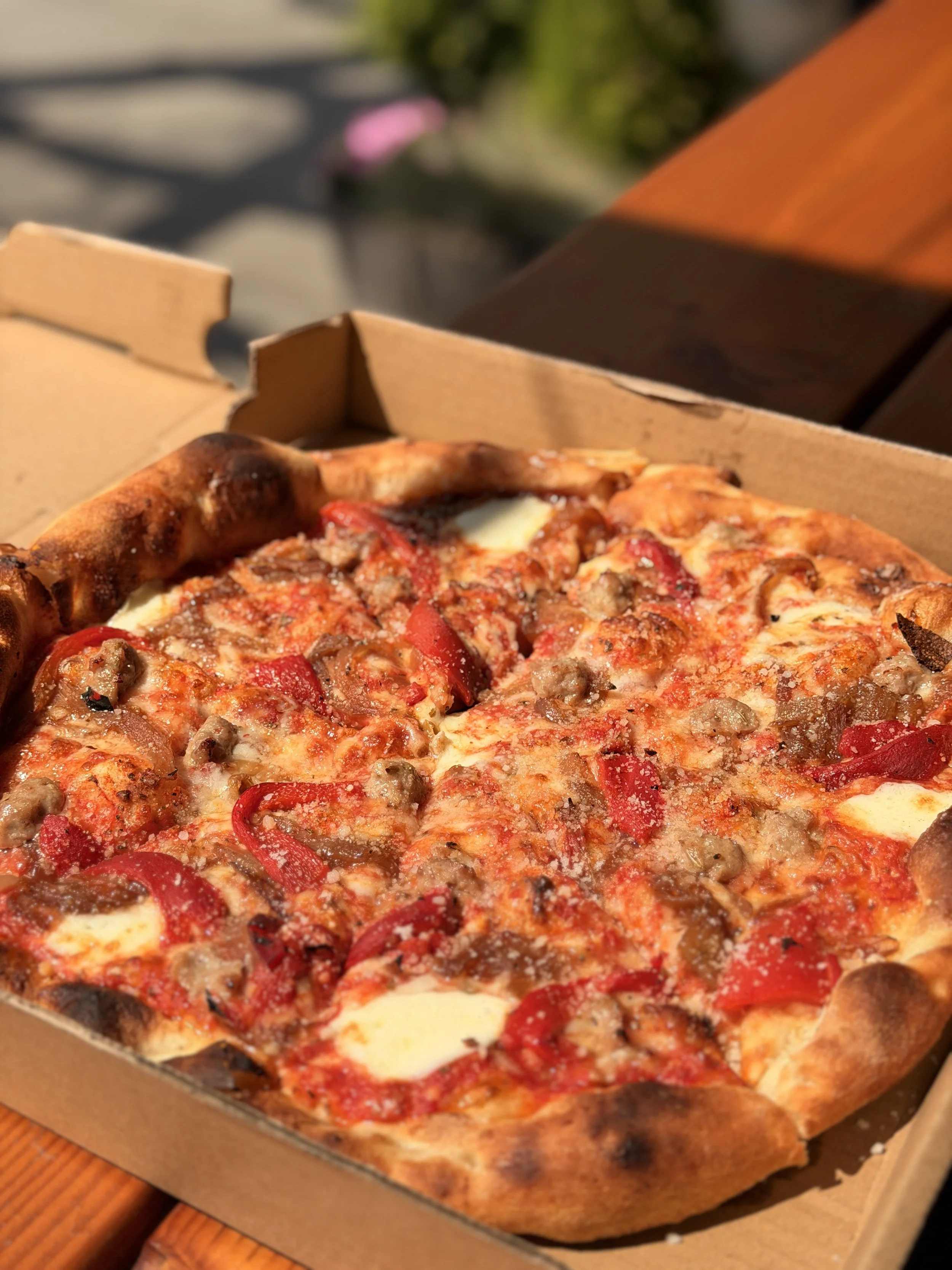 Close-up of a freshly baked pizza with pepperoni, sausage, and red peppers in a cardboard box.