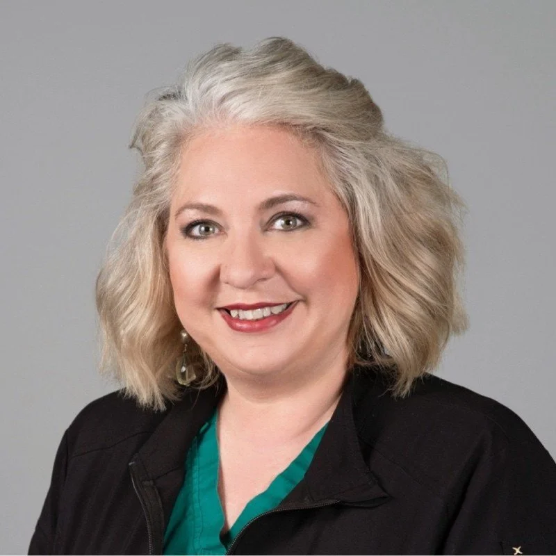 A woman with shoulder-length blonde curly hair smiling, wearing a black jacket and teal shirt against a gray background.