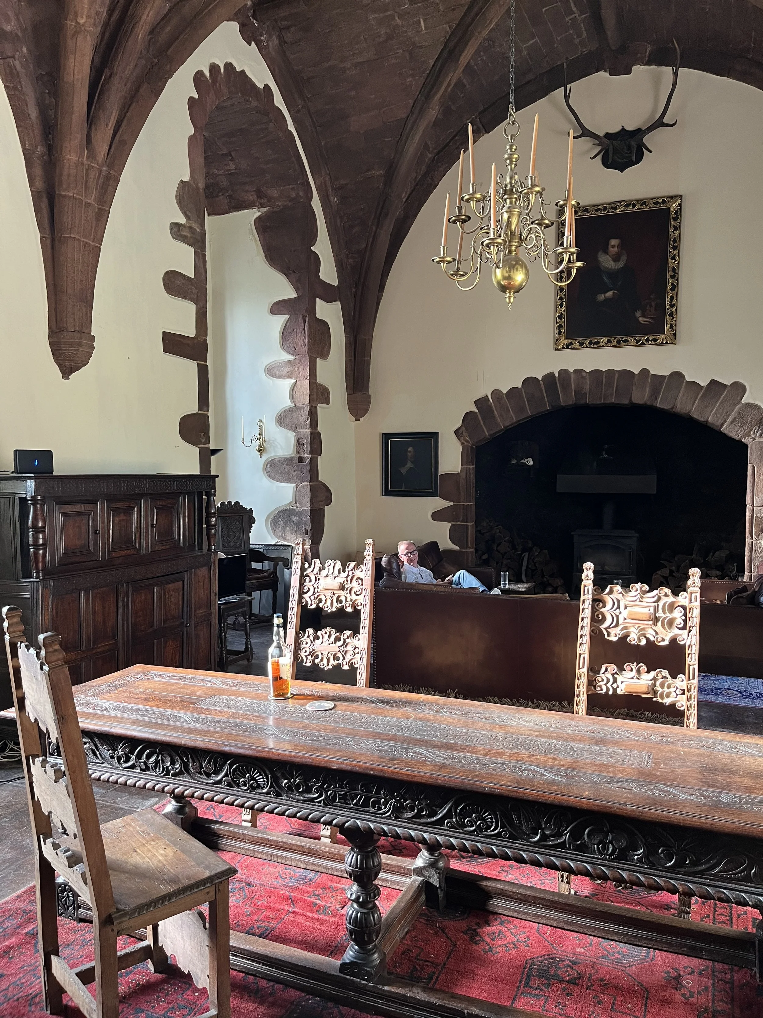 A large dining or sitting room with ornate wooden furniture, a red patterned rug, and high vaulted wooden ceilings with stone accents. There is a man sitting on a leather sofa, a large fireplace, and framed portraits on the wall. A chandelier hangs from the ceiling.
