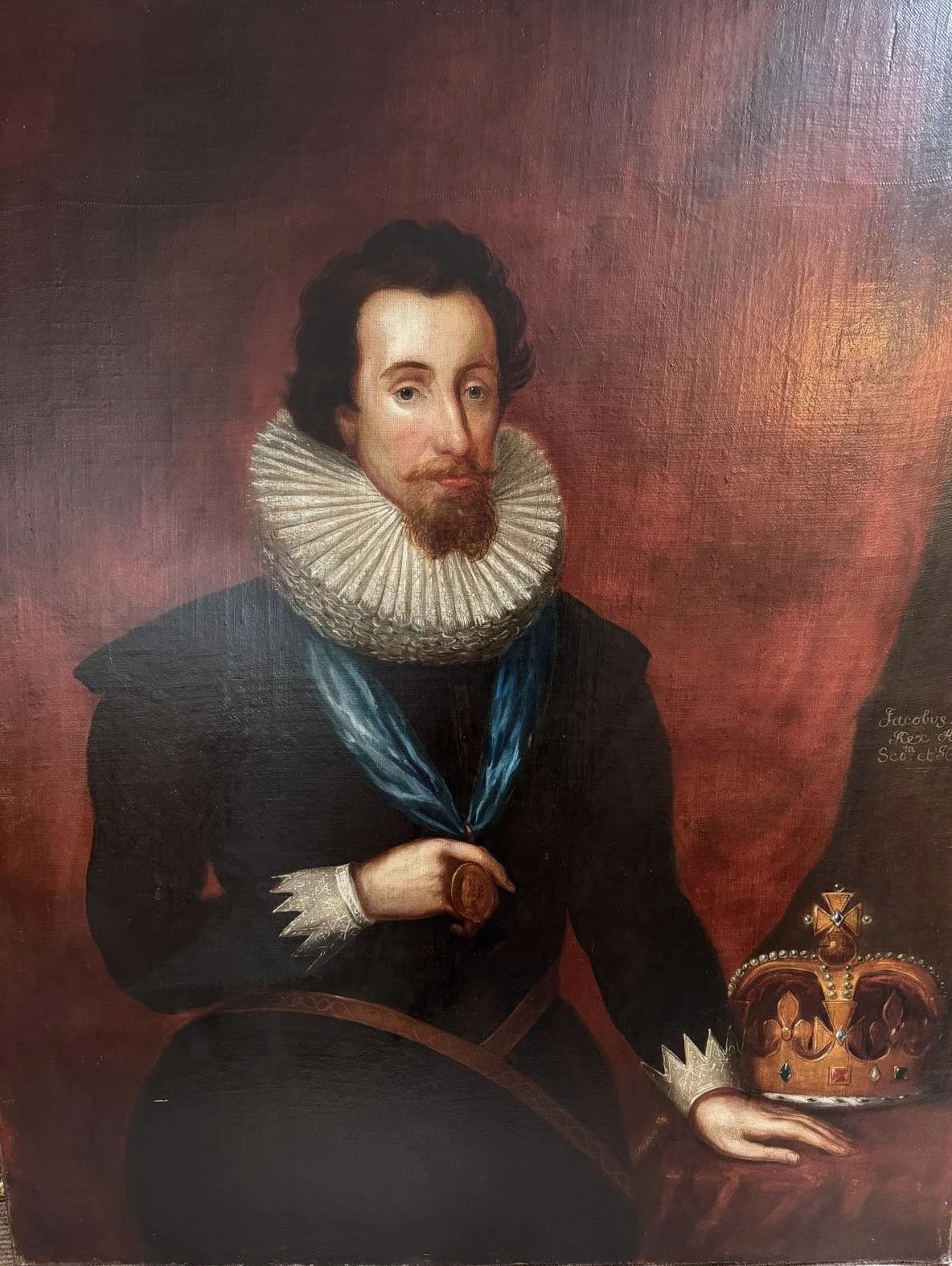 Portrait of King James VI and I, wearing a ruffled collar, holding a seal, seated next to a crown on a red background.