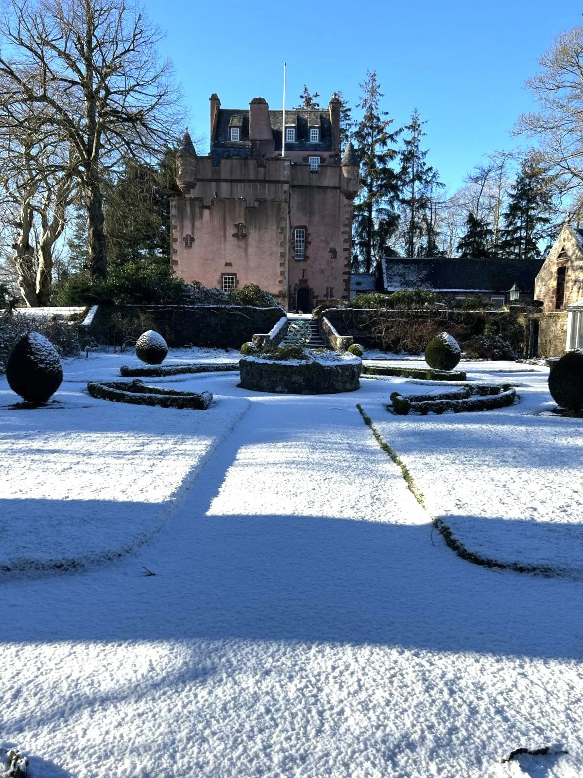 A snow-covered Towie Barclay Castle, surrounded by tall trees, with shadowed pathways and decorative bushes in the snow.