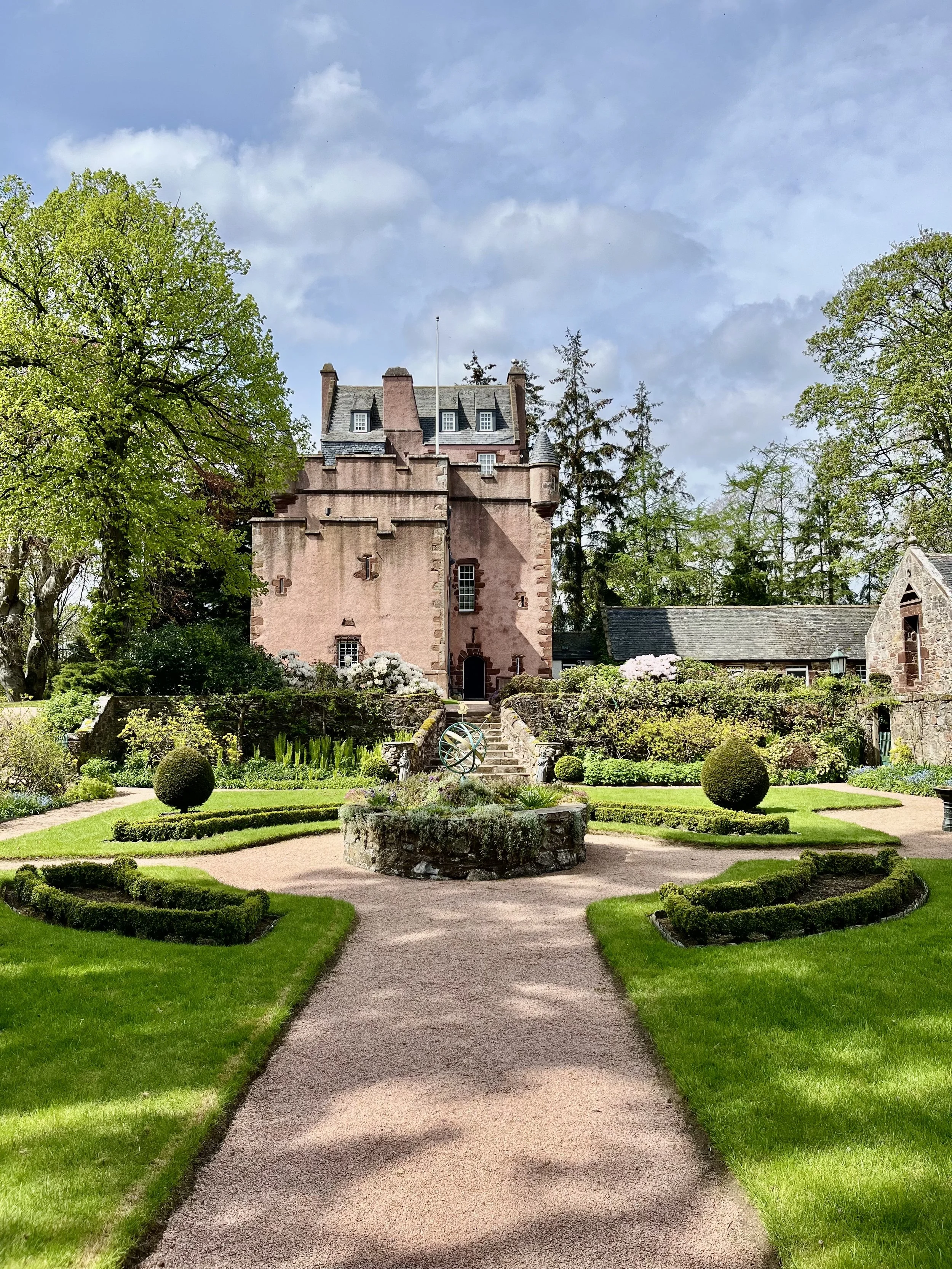 A stone pathway in a lush, green garden leads to a historic pink castle with multiple chimneys and turrets, surrounded by trees and flowering shrubs under a partly cloudy sky.