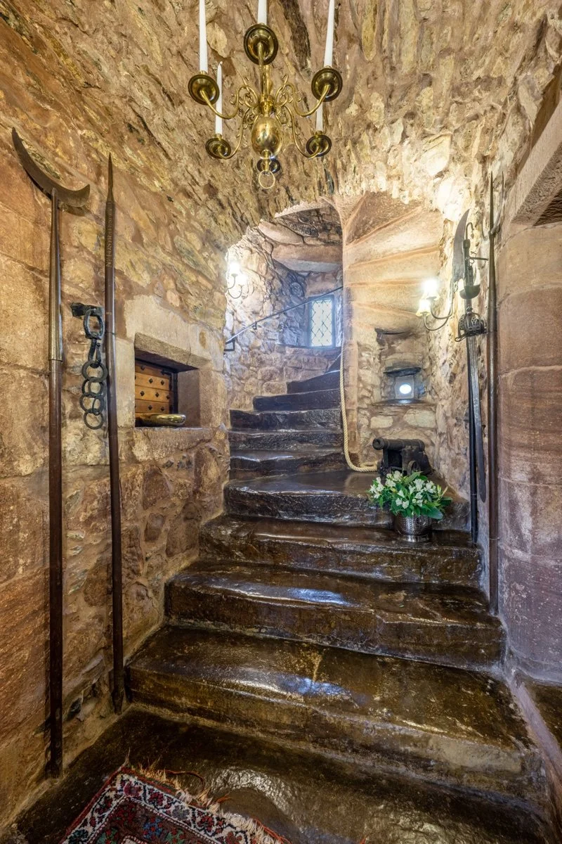 Stone staircase in a castle or historic building, with medieval weapons and a chandelier hanging from a stone ceiling.