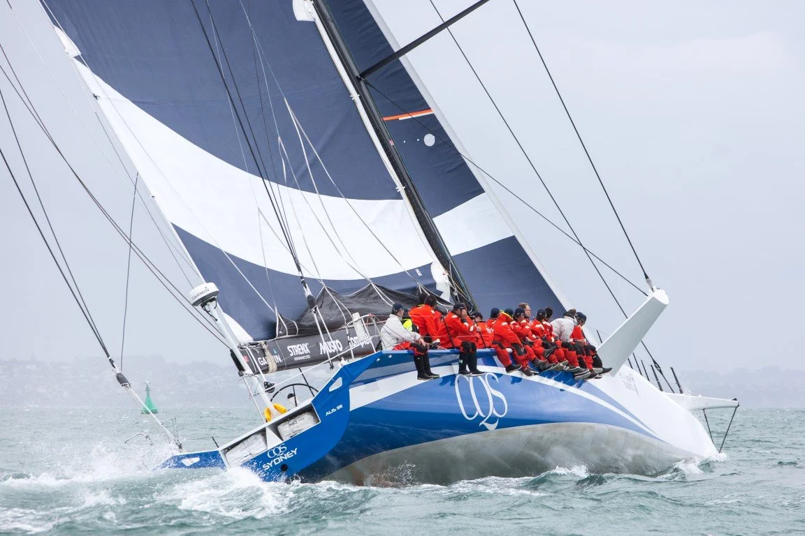 A large racing sailboat with numerous crew members wearing red jackets, sailing on choppy waters with its sails fully deployed.