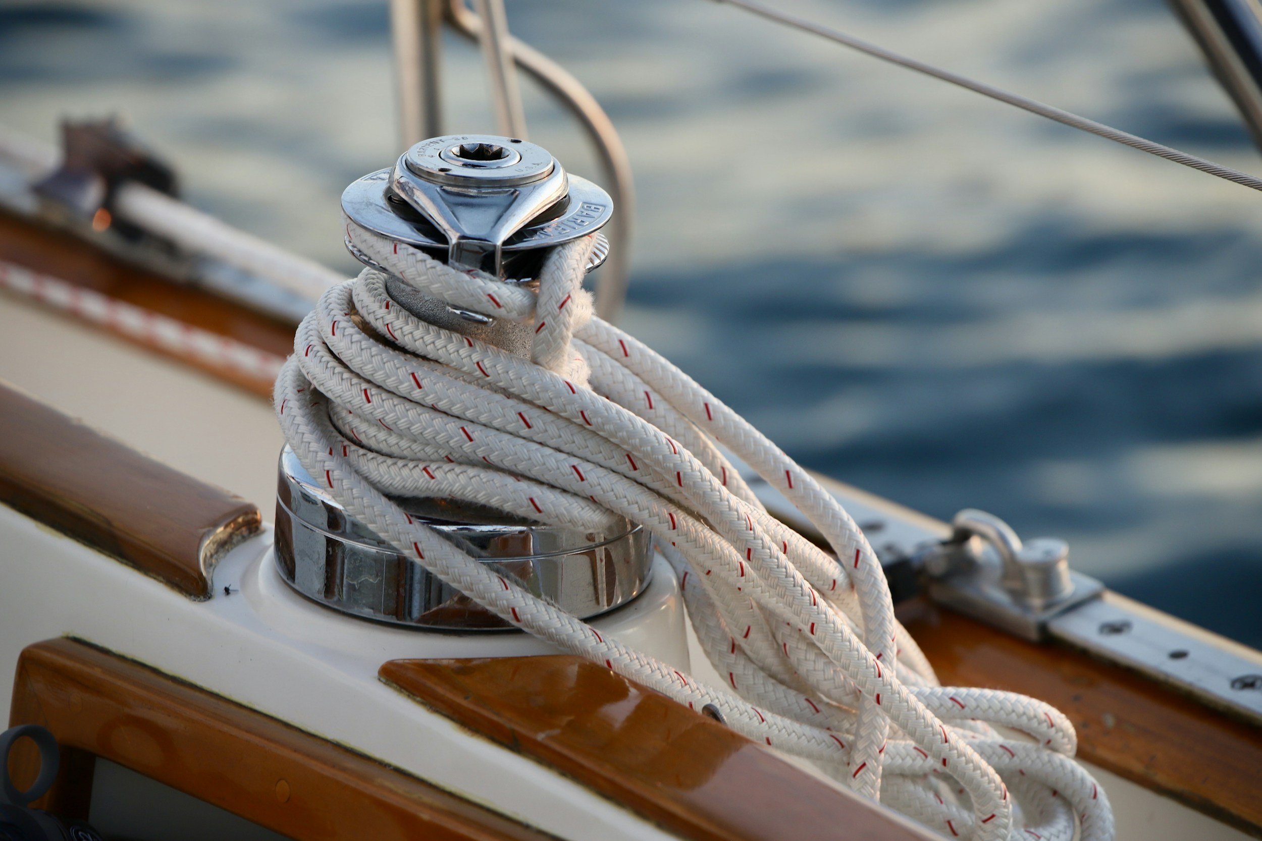 Close-up of a boat's metal winch with a white rope wrapped around it, set against a blurred background of water.