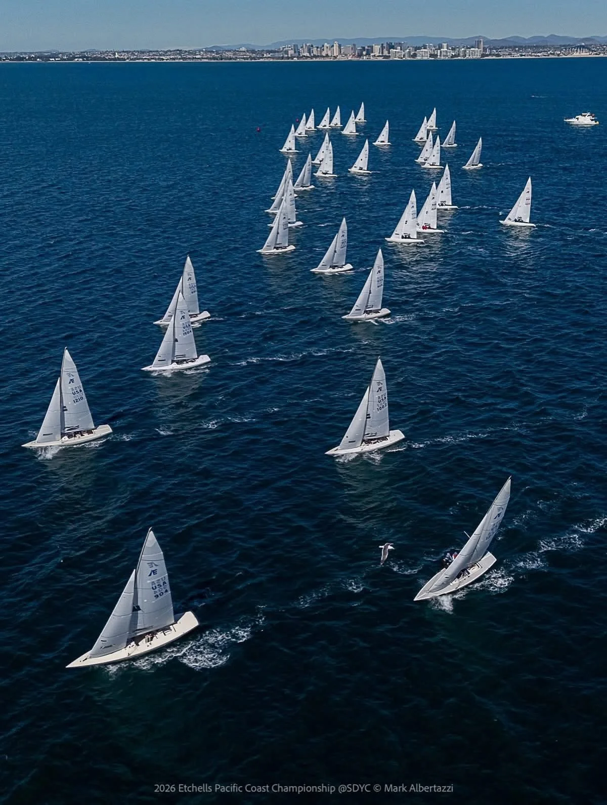 Several sailboats participating in a yacht race on the ocean near a city with high-rise buildings, with mountains in the background.