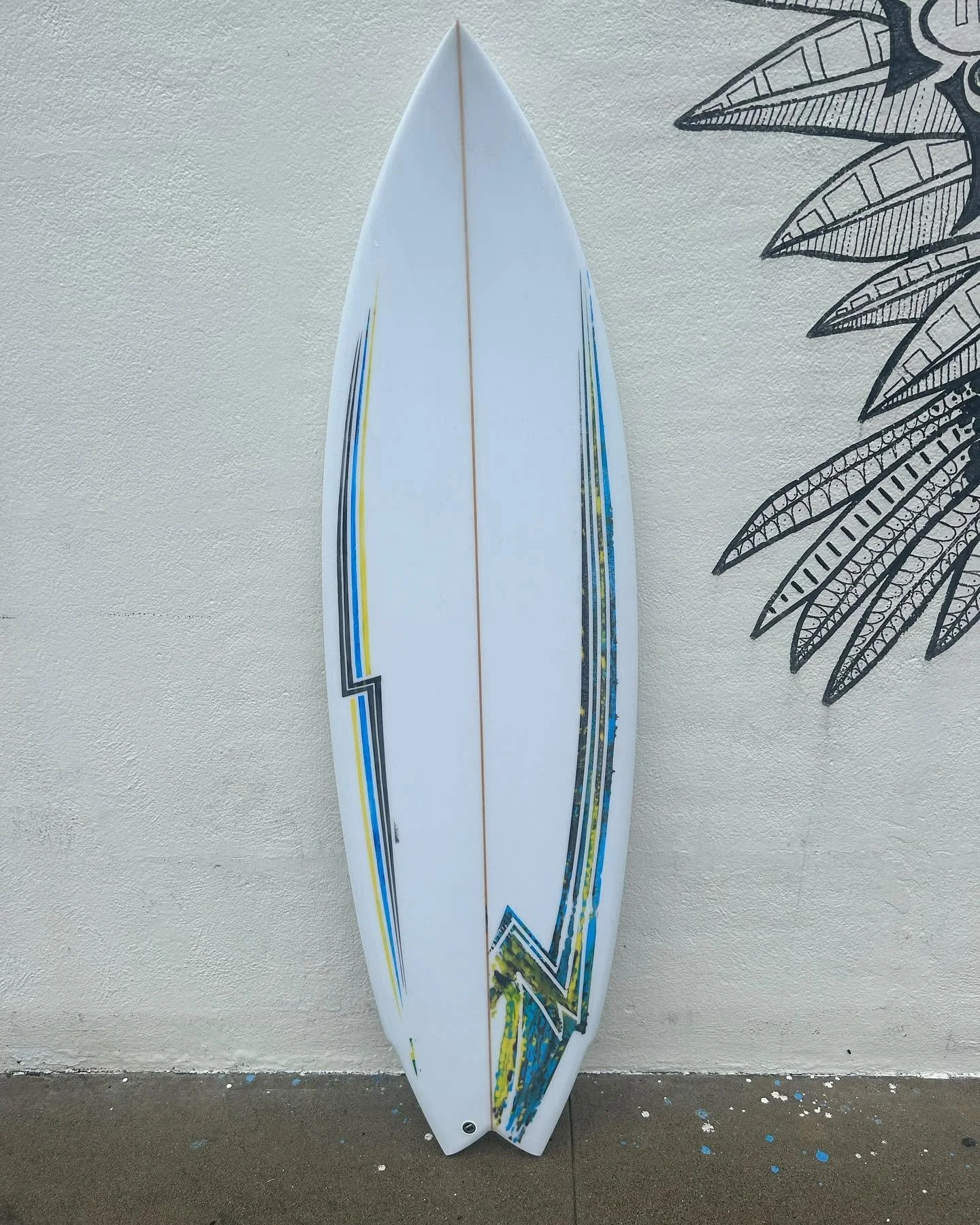 A white surfboard with colorful geometric designs leaning against a textured white wall with black leaf illustrations.