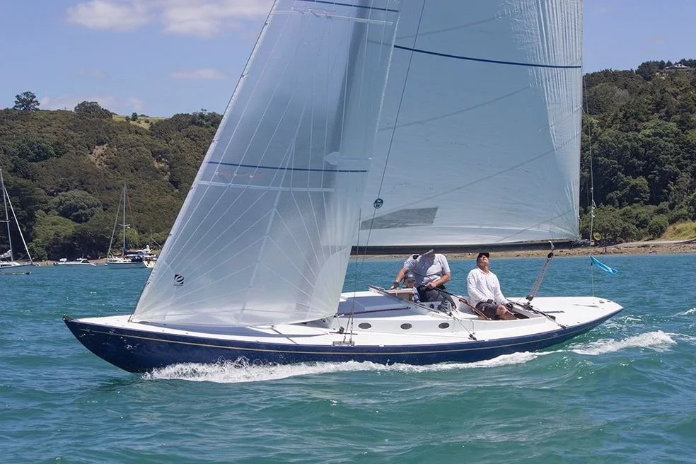 Sailboat with two people on board, sailing on a lake with hilly, wooded shoreline in the background.