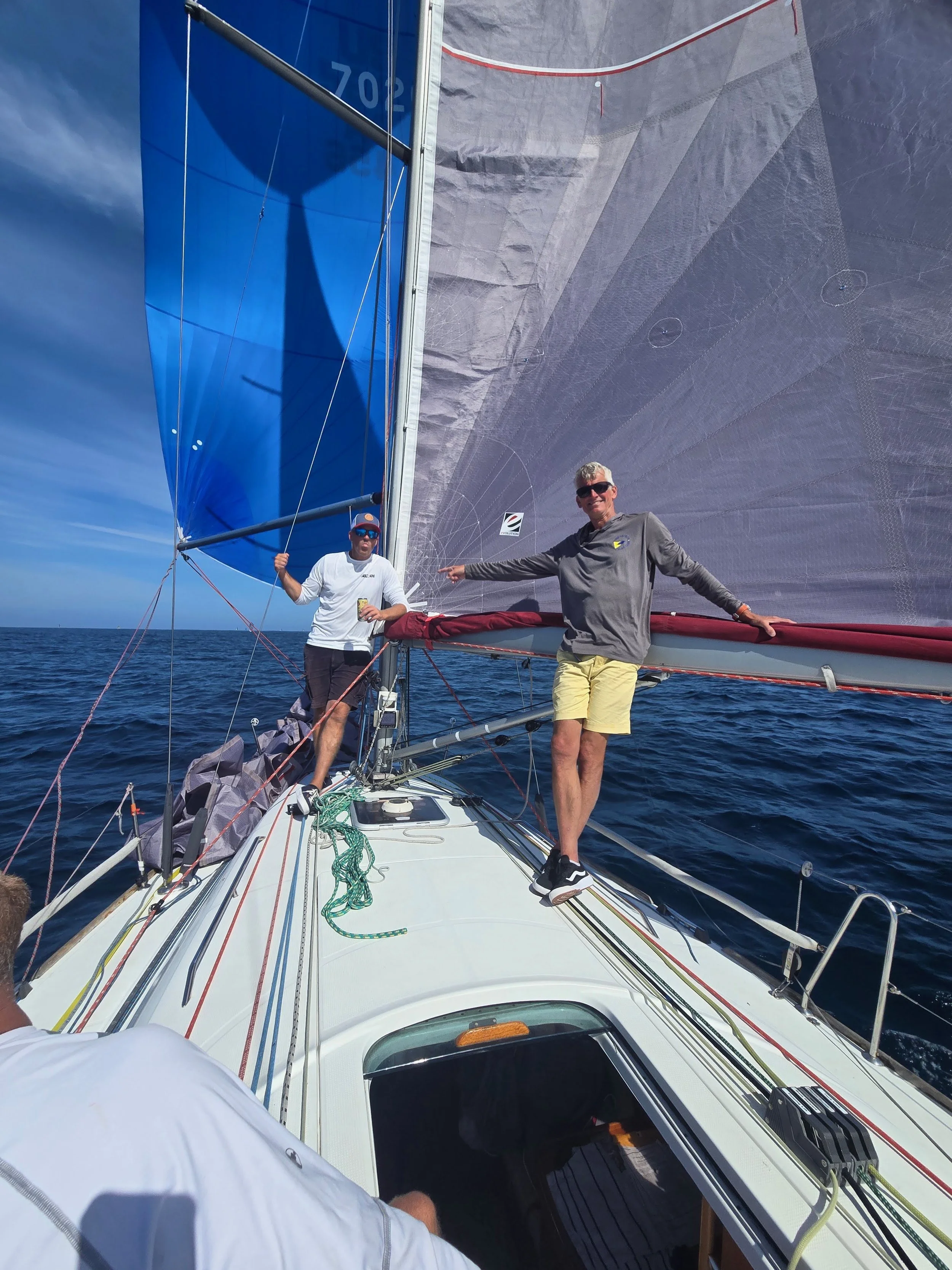 Two men standing on a sailboat, with a large sail behind them, on open water under a clear sky.