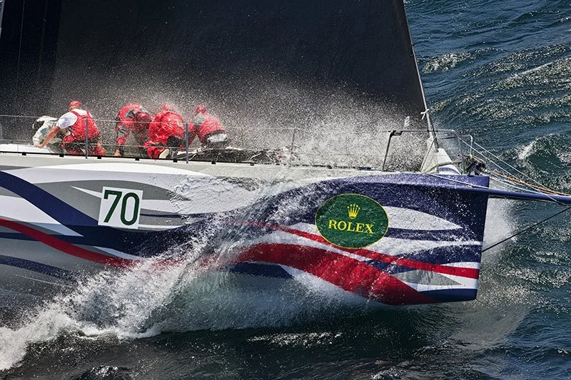 A racing sailboat with crew members in red suits, cutting through the water at high speed. The boat displays a Rolex logo and the number 70, with a black sail and colorful hull markings.