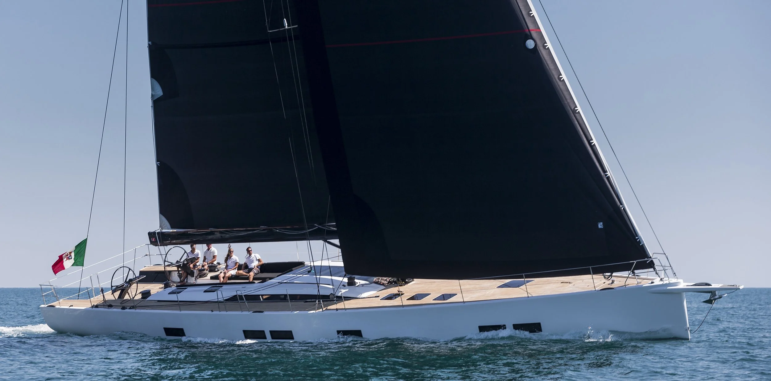 A sailing yacht with a black sail on the water, carrying four people on the deck, with an Italian flag at the stern, during daytime.