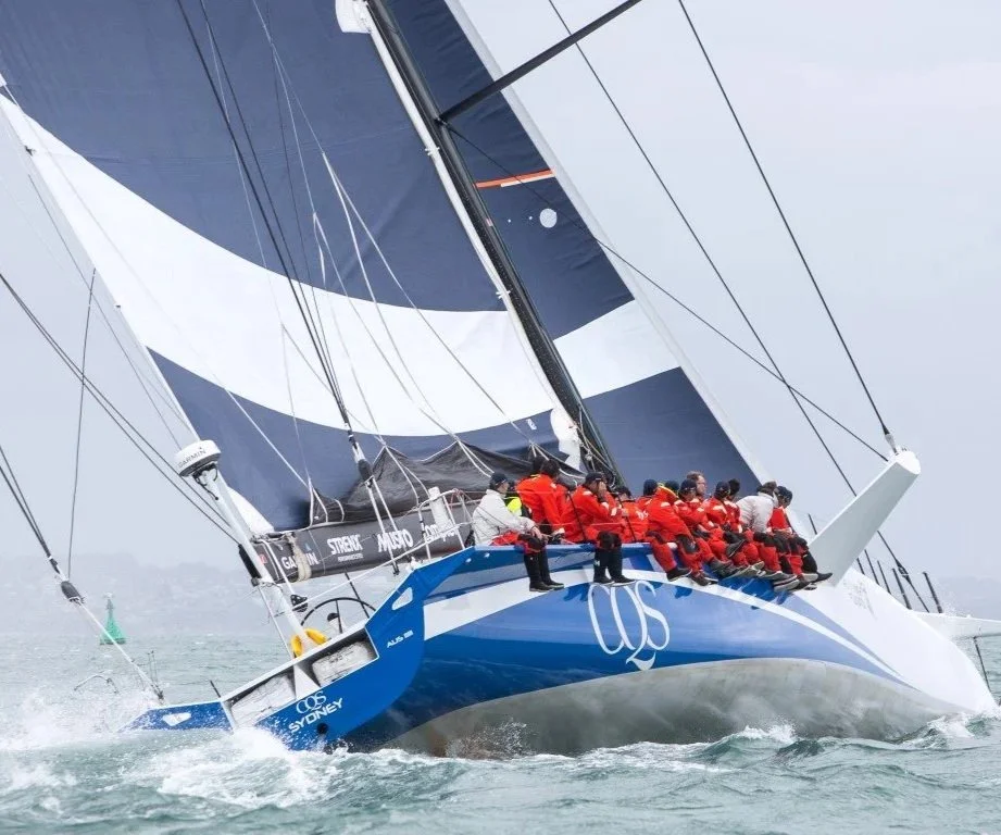 A sailboat with multiple crew members wearing red jackets racing on the water, with large sails in a steep angle.