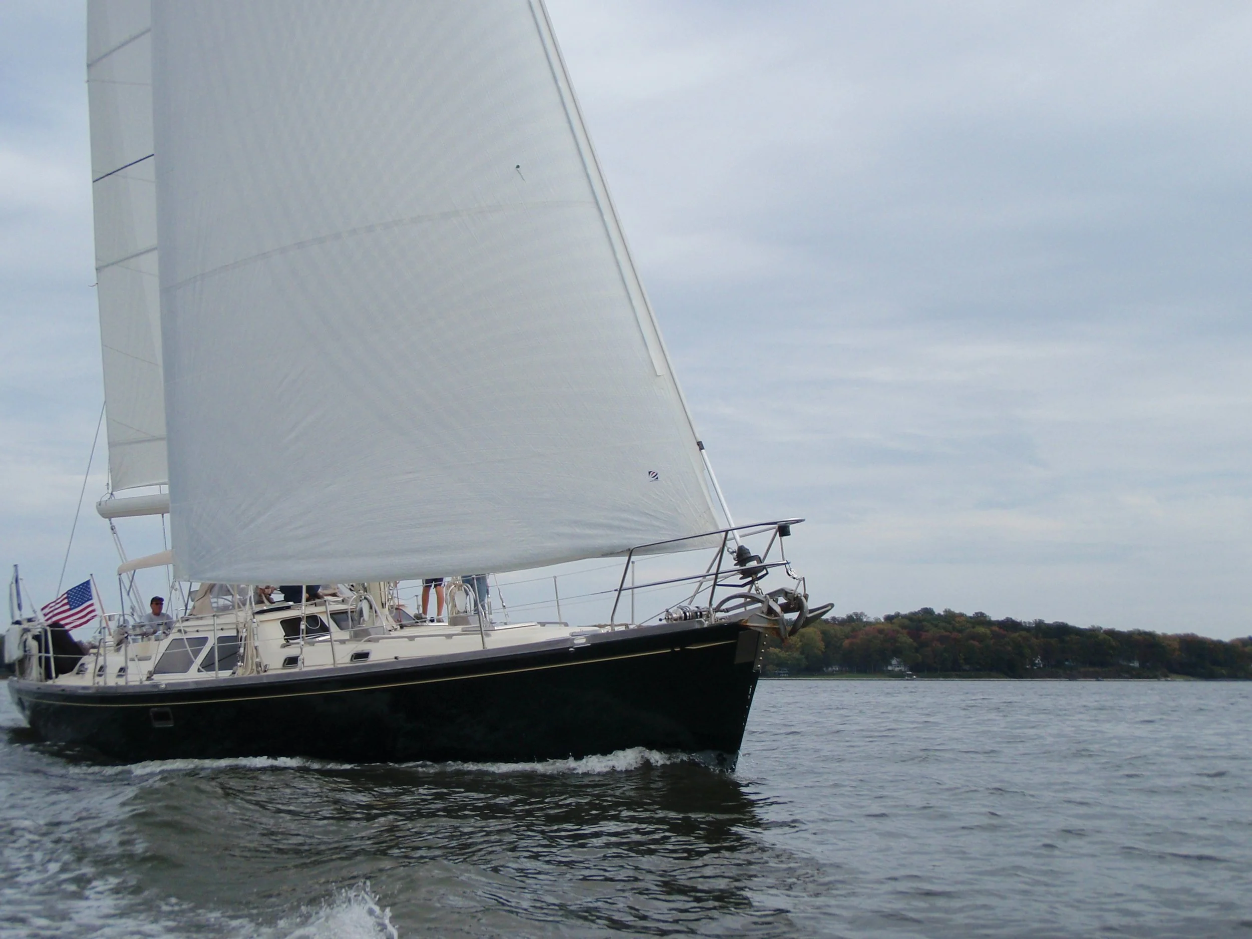 A sailboat with white sails sailing on a body of water with trees in the background and at least one person on board.