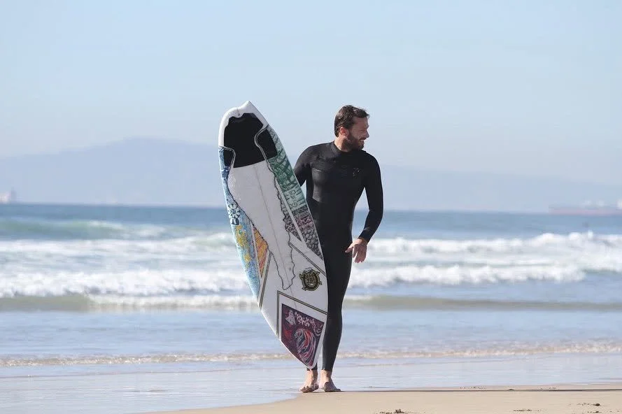 Man in a wetsuit holding a surfboard walking on the beach near the ocean with waves and distant ships.