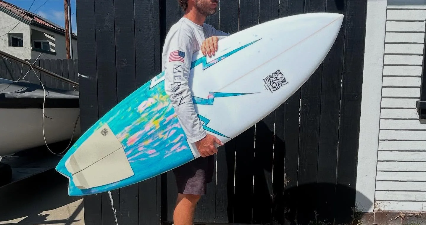 Person holding a colorful surfboard outside next to a black wooden fence.