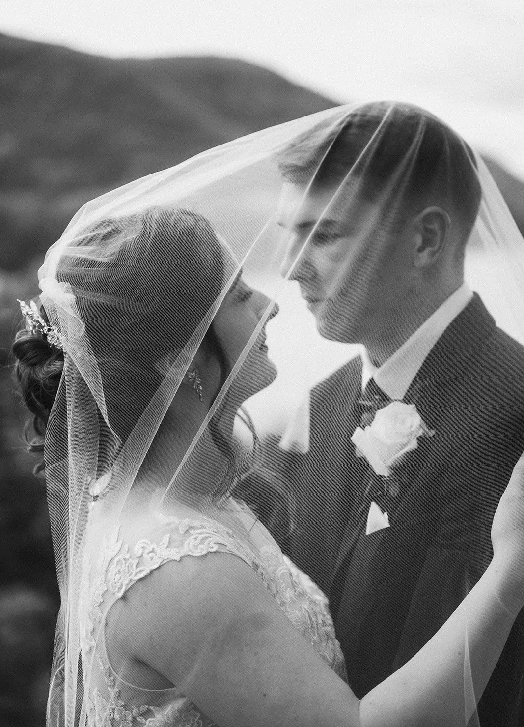 Black-and-white photo of a bride and groom under a veil, facing each other with mountain in background.