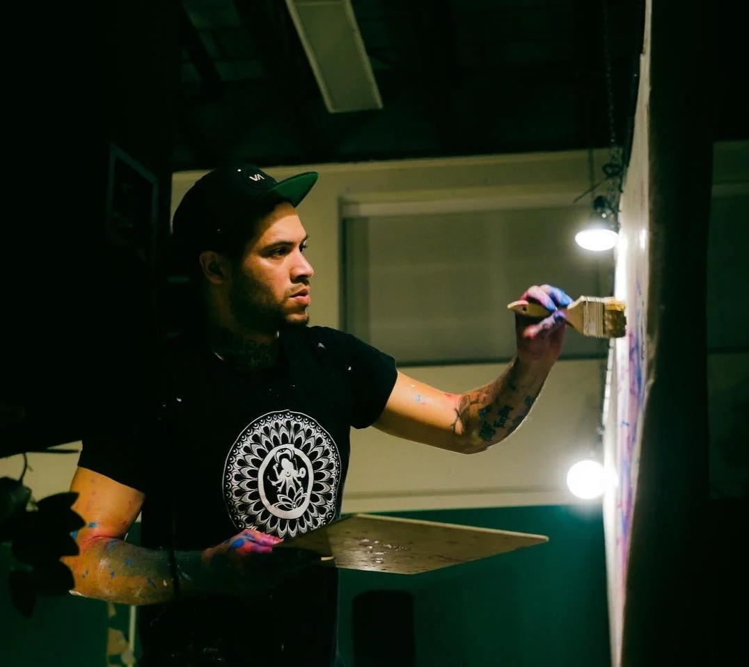 A man with tattoos and a black cap working on an art piece on a wall, using a paintbrush under a bright overhead light.