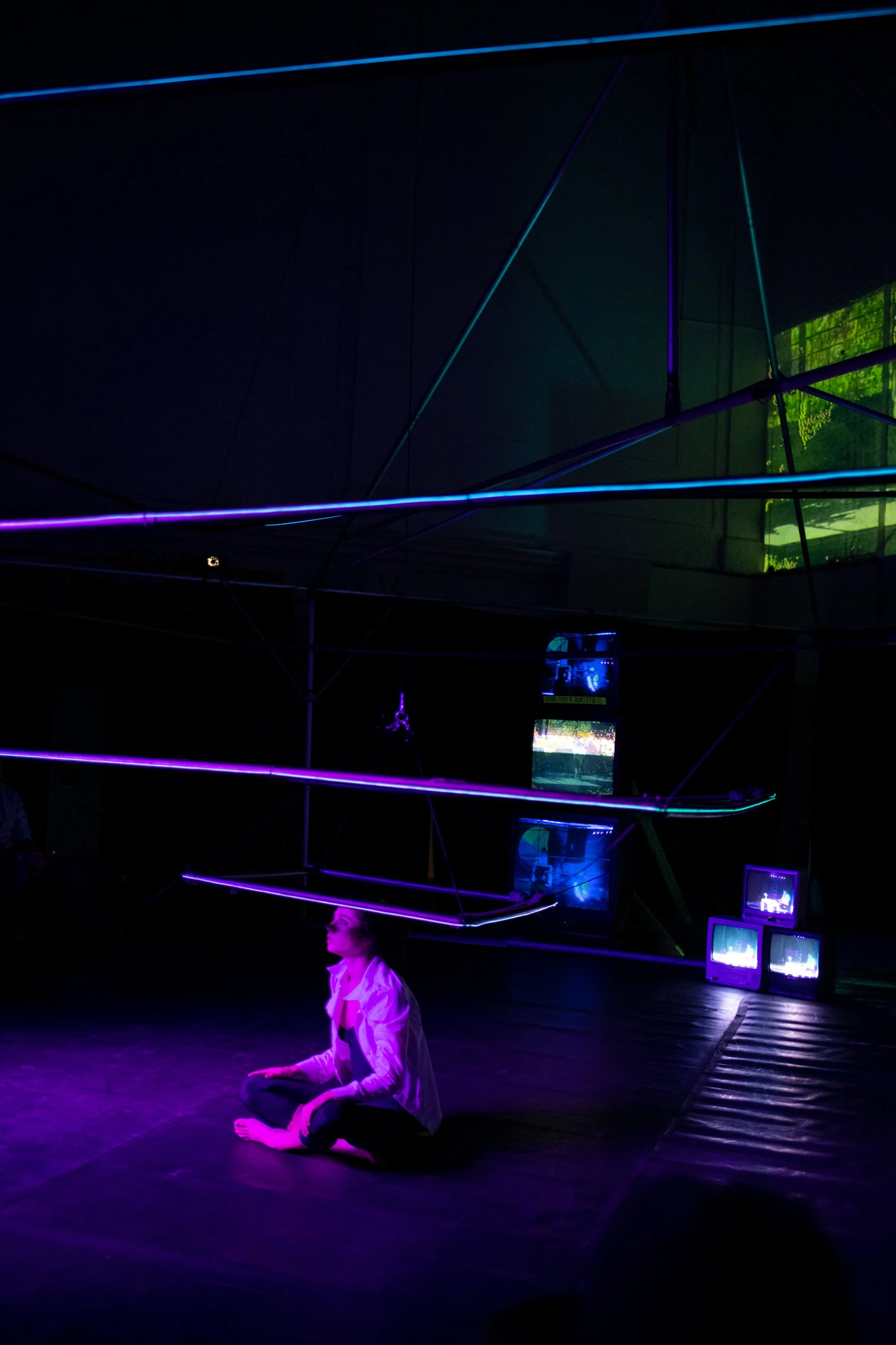 A woman in white clothing sitting on the floor in a dark room with neon lights and old television screens.